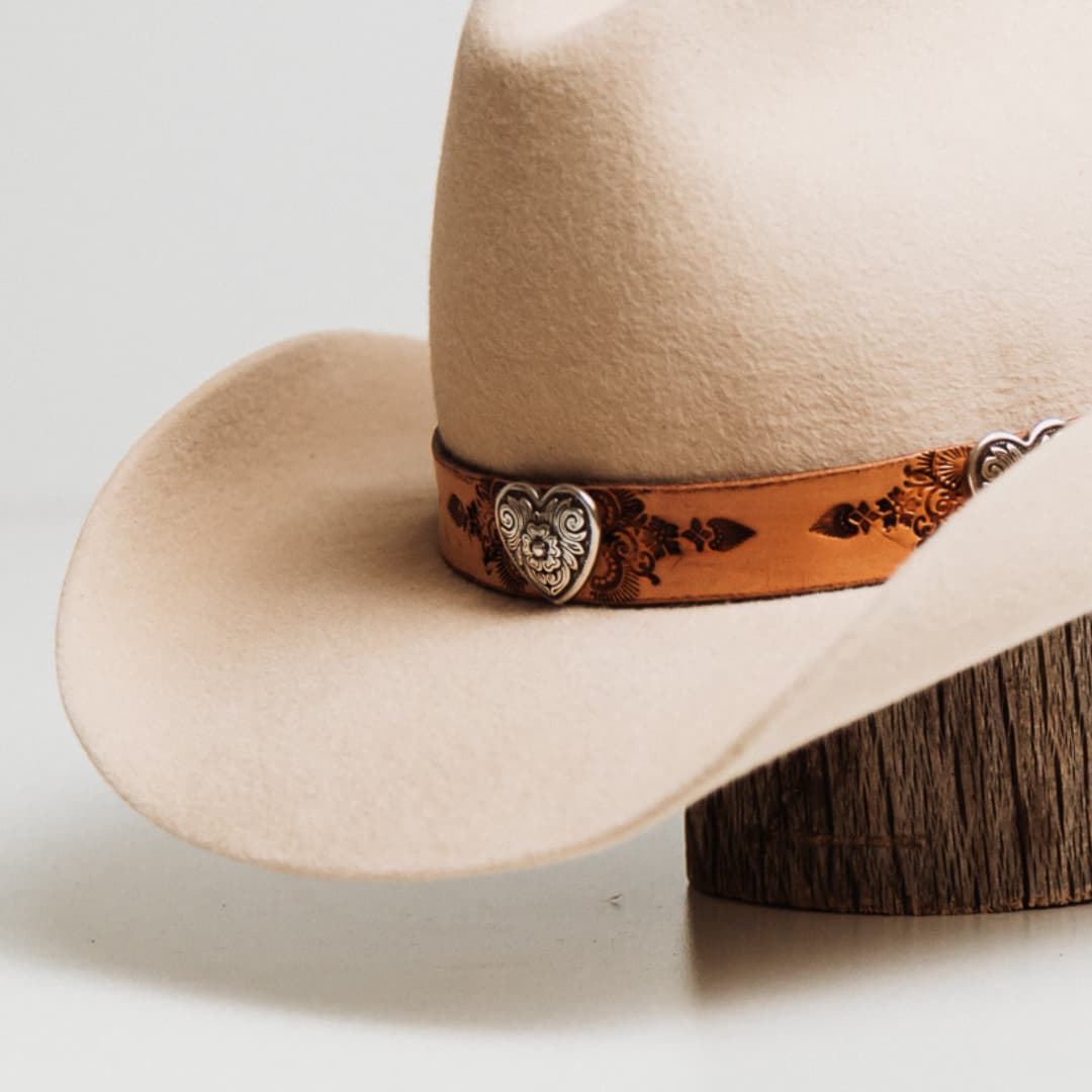 Close up detail of the Phylli x Buffalo Girl Palamino Heart Concho Hat Band showing silver plated heart concho set on hand tooled Palamino leather Mandala hat band on a beige felt cowboy hat by PHYLLi Designs.