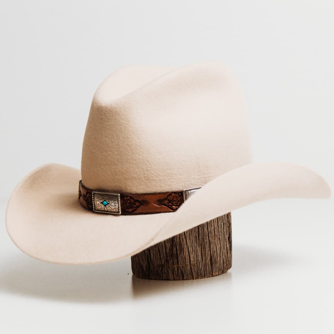 Front left angle of the PHYLLi x Buffalo Girl Rugged Desert Buckle Hat Band with antique brown hand tooled leather hat band and turquoise conchos on a beige felt cowboy hat by PHYLLi Designs.