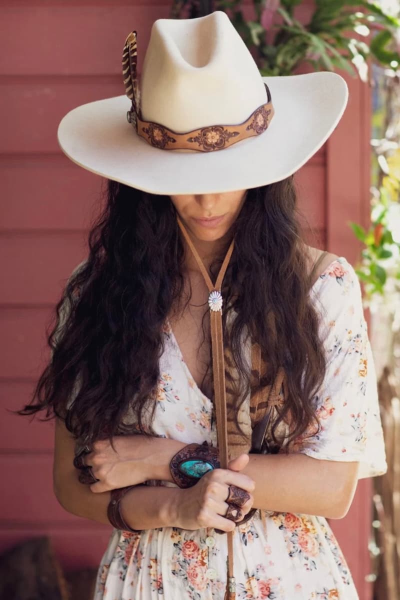 Woman wearing the PHYLLi x Buffalo Girl Stampede Strap attached to a felt hat, holding the strap beneath her chin while standing outdoors in a garden setting.