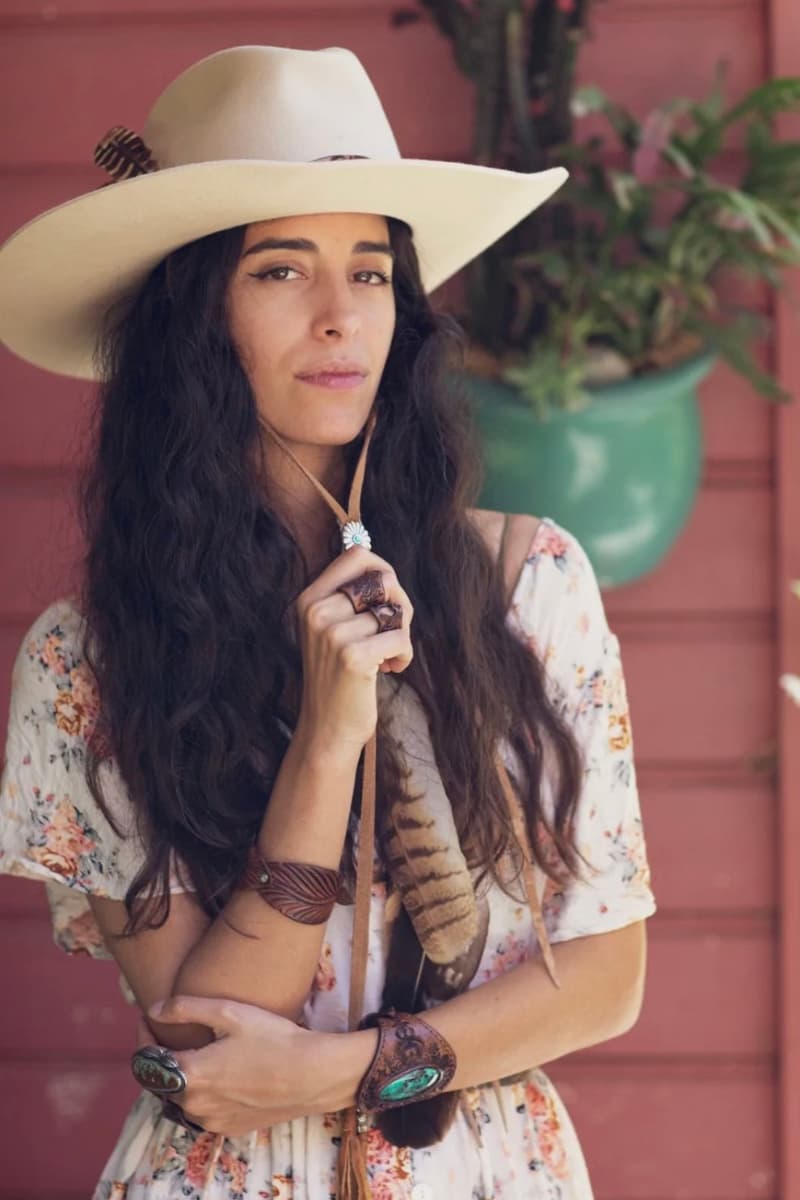Woman wearing a felt hat fitted with the PHYLLi x Buffalo Girl Stampede Strap in tan suede while standing outdoors against a rustic backdrop.