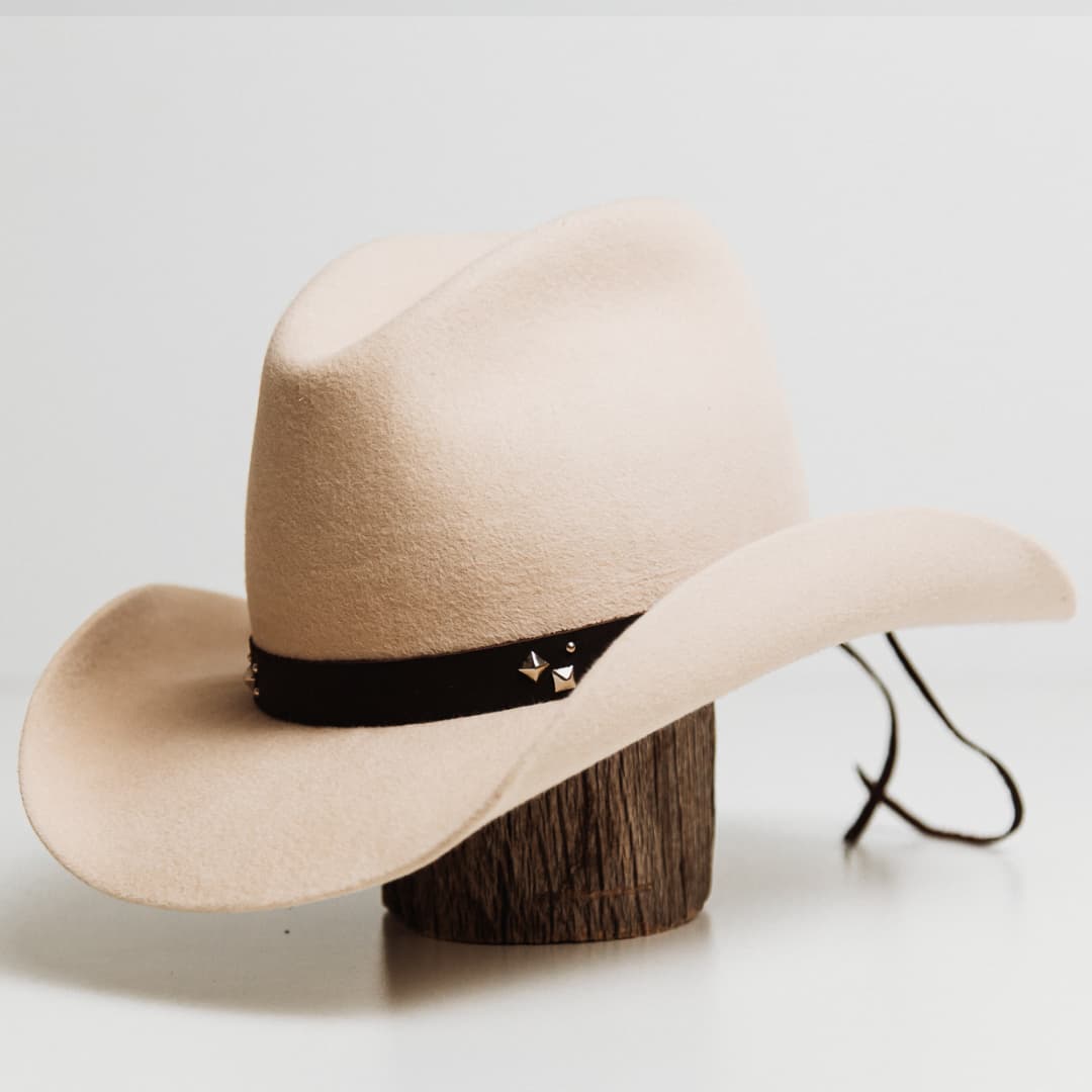 Front left angle of the Buffalo Tieback Studded Hat Band featuring dark brown buffalo hide band with stainless steel studded detailing on a beige felt cowboy hat by PHYLLi Designs.