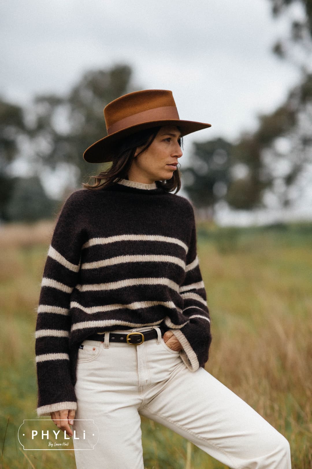 Woman wearing the Calaveras caramel felt hat by PHYLLi Designs with chocolate grosgrain band and suede string in open countryside grasslands.