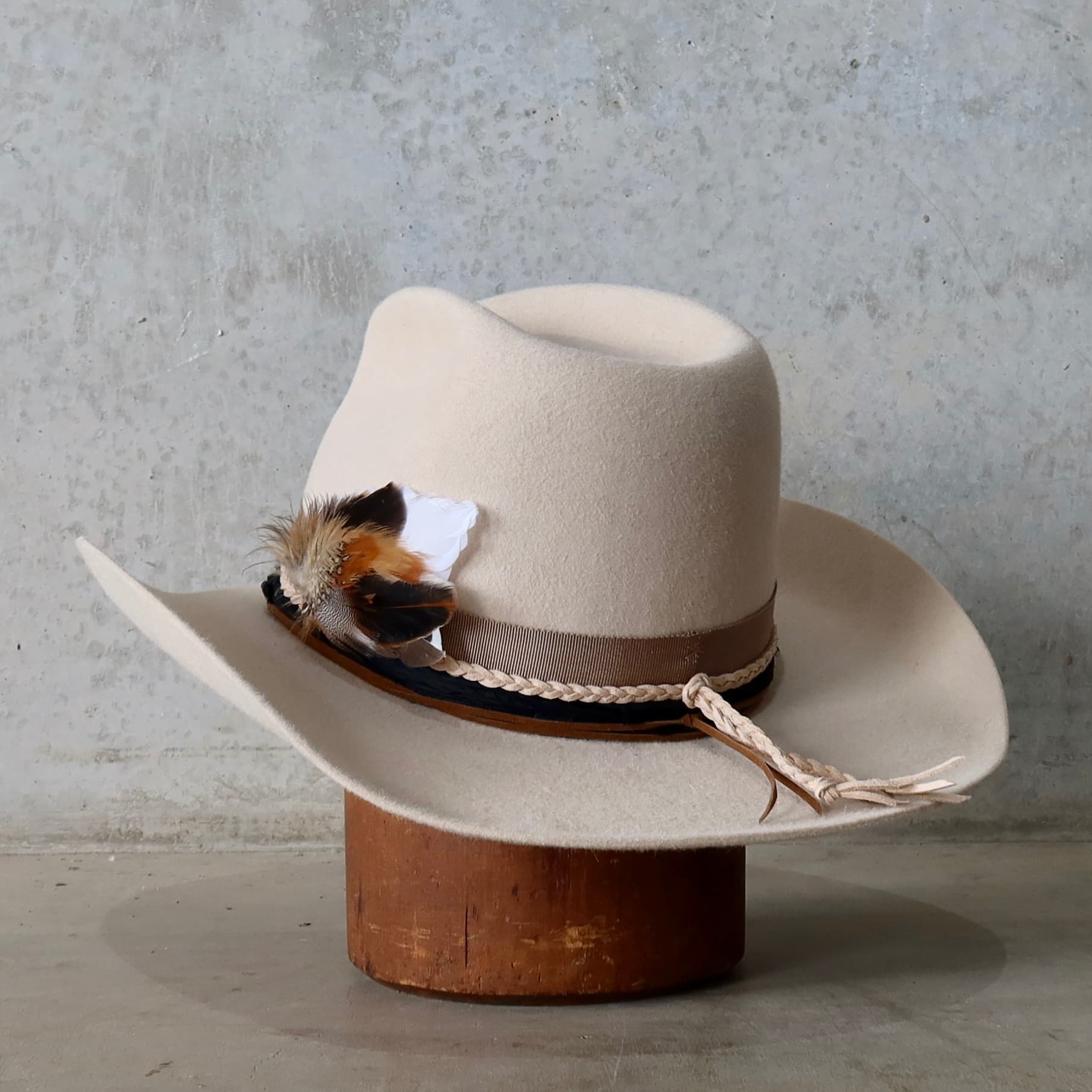 Front left angle of the beige felt cowboy hat Casey Beige by PHYLLi Designs with taupe grosgrain band, plaited nude suede string and fallen feather bunch.