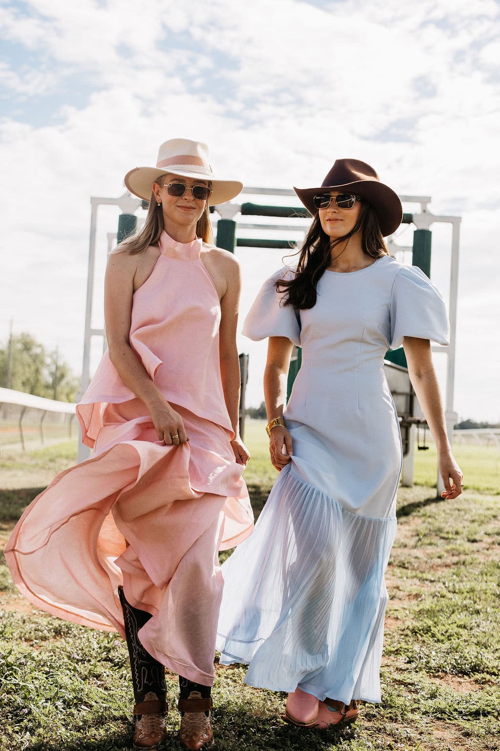 Woman wearing the PHYLLi Clementine beige wide brim felt hat and a flowing dress, standing with a friend on grass by the starting gate.