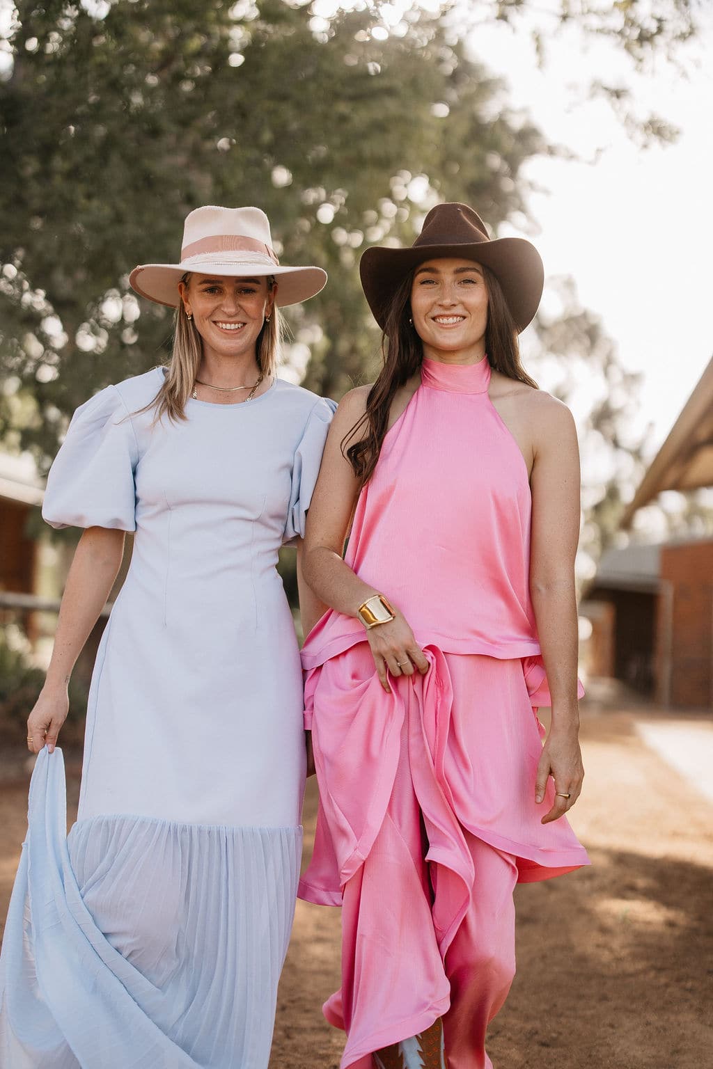Two women walking outdoors amon trees, one wearing the PHYLLi Clementine beige wide brim felt hat with a pale blue dress.