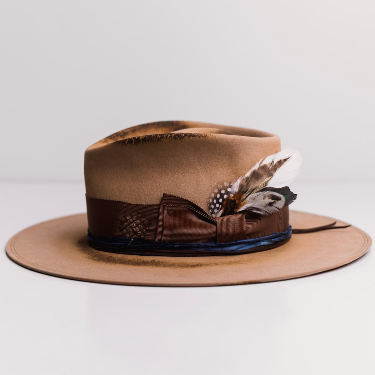 Left side view of the Columbian camel felt hat by PHYLLi Designs with chocolate grosgrain bow band, azure sari silk trim and feather detail.