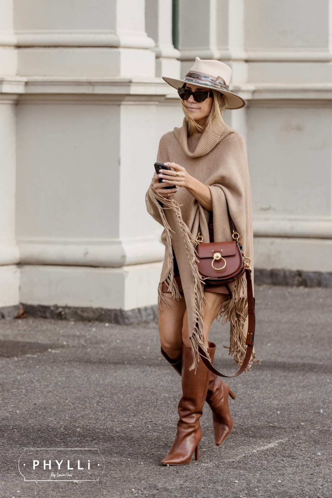 Woman wearing the Cunningham beige felt hat by PHYLLi Designs with taupe grosgrain band and sari silk trim while walking outdoors beside a stone building.