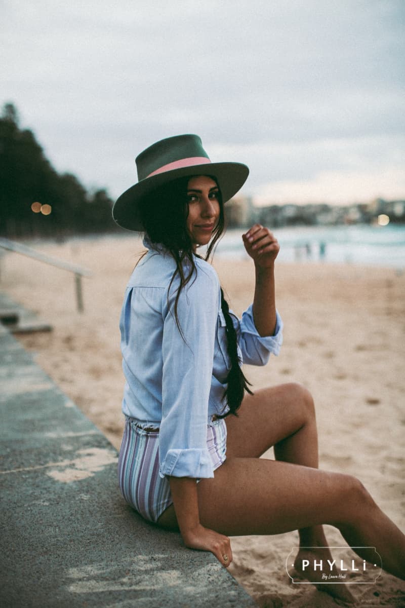 Woman wearing the Diamonds & Pearls eucalyptus felt hat by PHYLLi Designs with sherbet pink grosgrain band and cream sari silk trim on a beach.