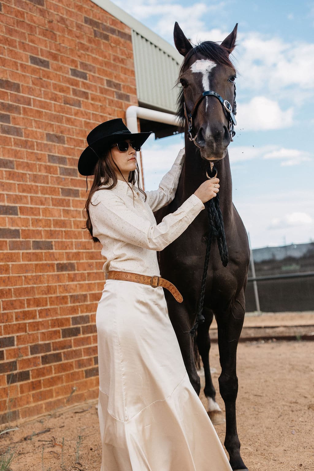 Woman wearing the PHYLLi Edwards black felt cowboy hat with a cream silk shirt and skirt, standing beside a horse outside.