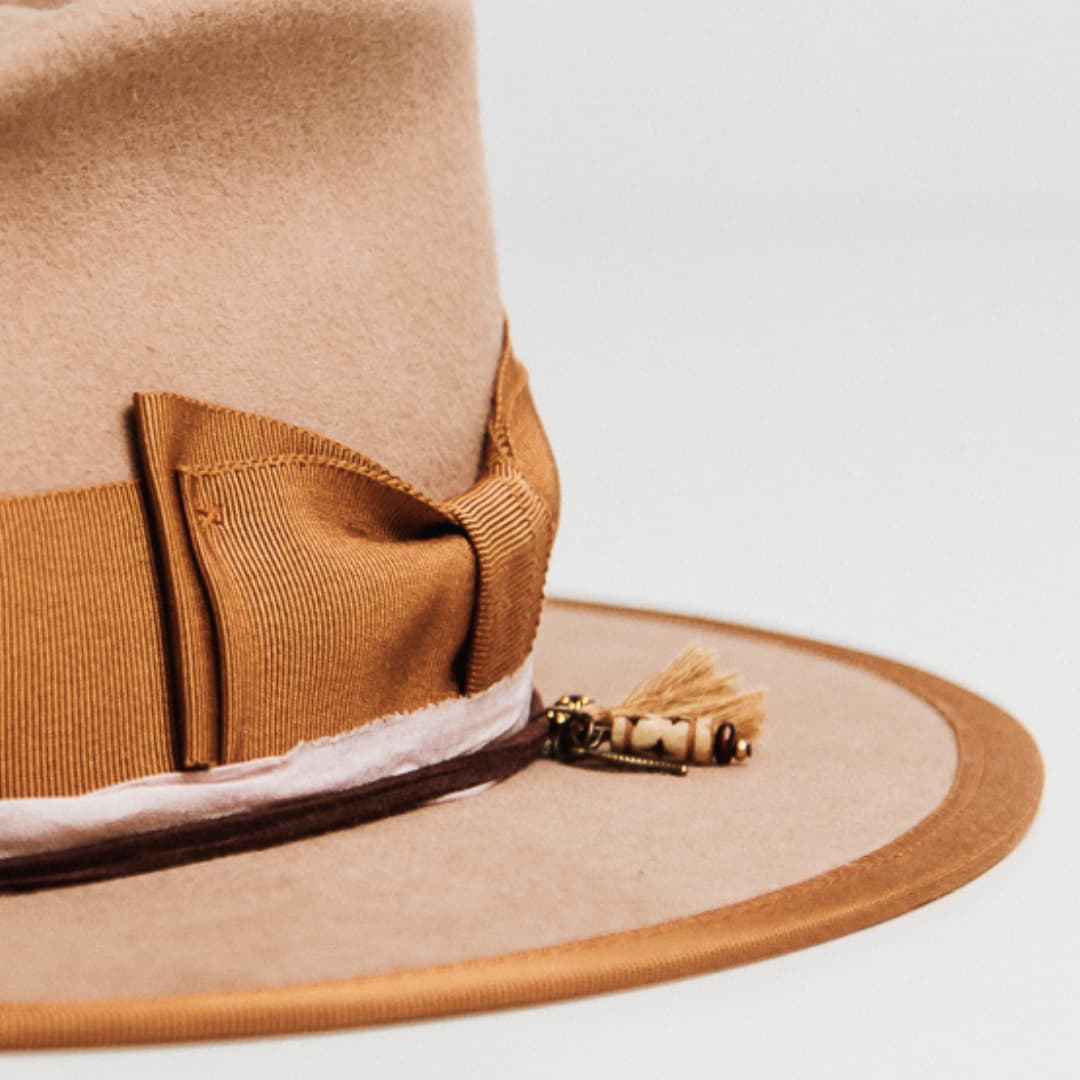 Detail of Feuille Morte Camel felt hat showing Copper grosgrain double bow band and charm cluster with tassel, bronze leaf charm and timber bead.