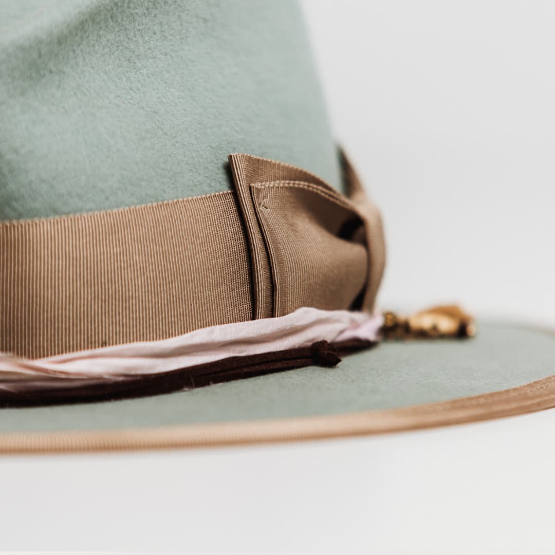 Detail of Feuille Morte Sage felt hat showing taupe grosgrain double bow band and charm cluster with tassel, bronze leaf charm and timber bead.