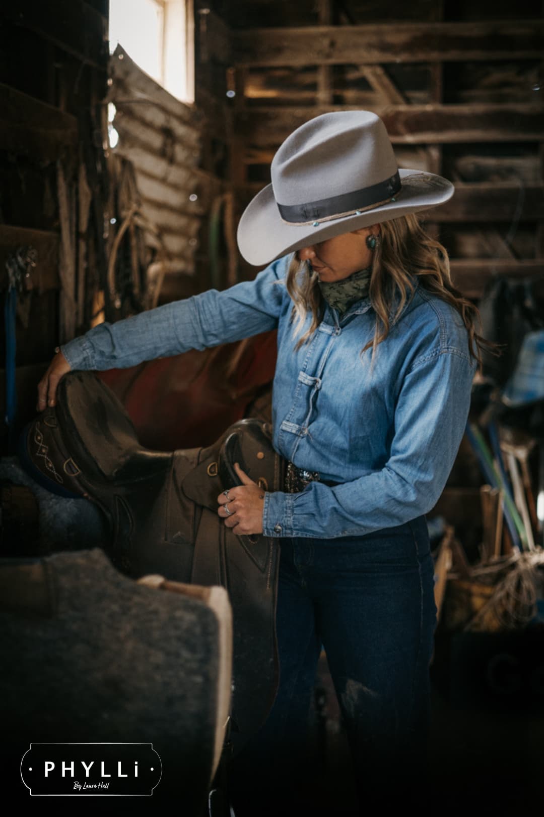 Model wearing the Finse cement felt cowboy hat by PHYLLi Designs with turquoise beaded twine wraps holding a saddle in a horse stable.