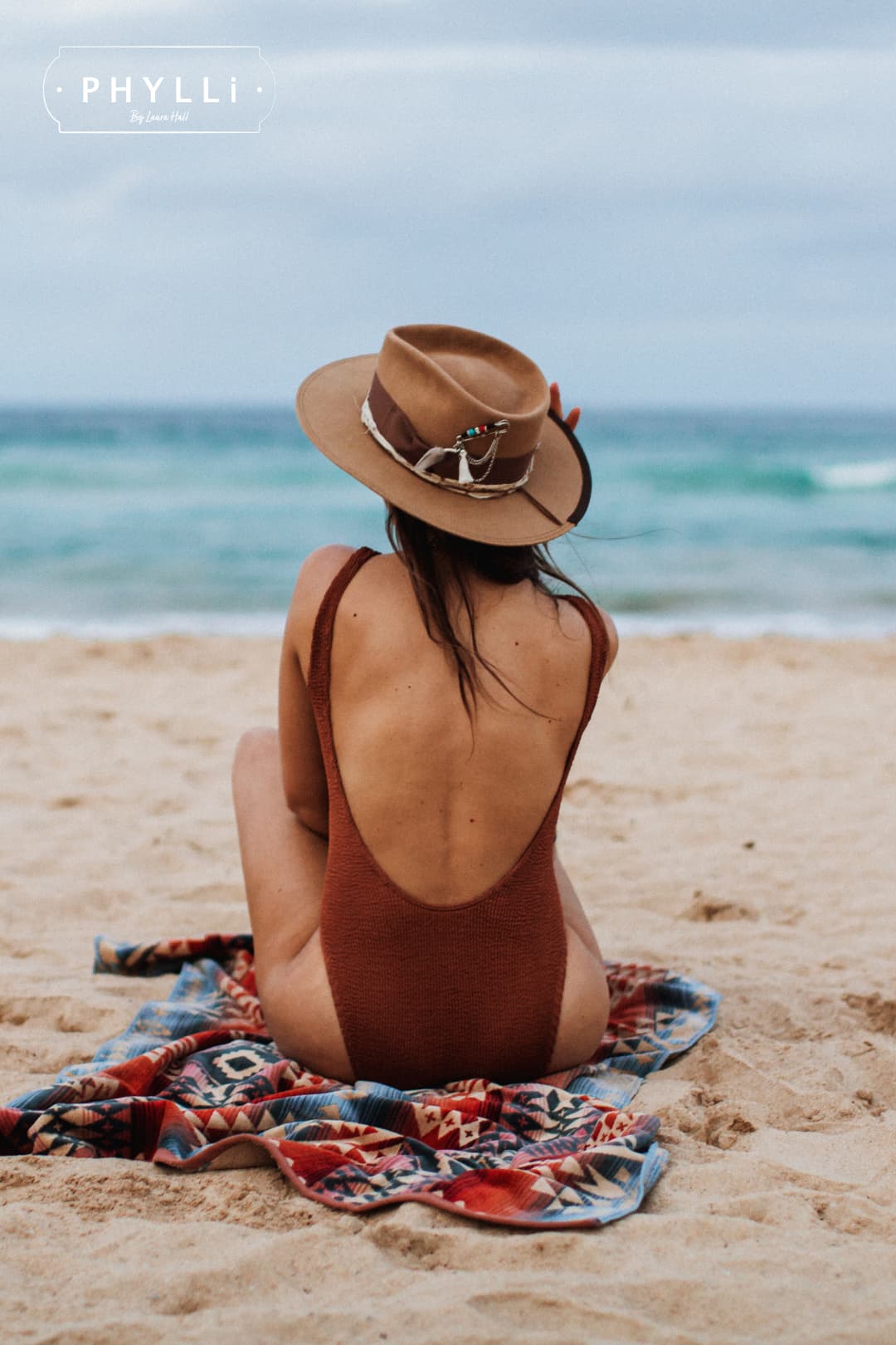 Back view of woman wearing Fly Fisher camel felt hat by PHYLLi Designs while sitting on a beach towel facing the ocean.