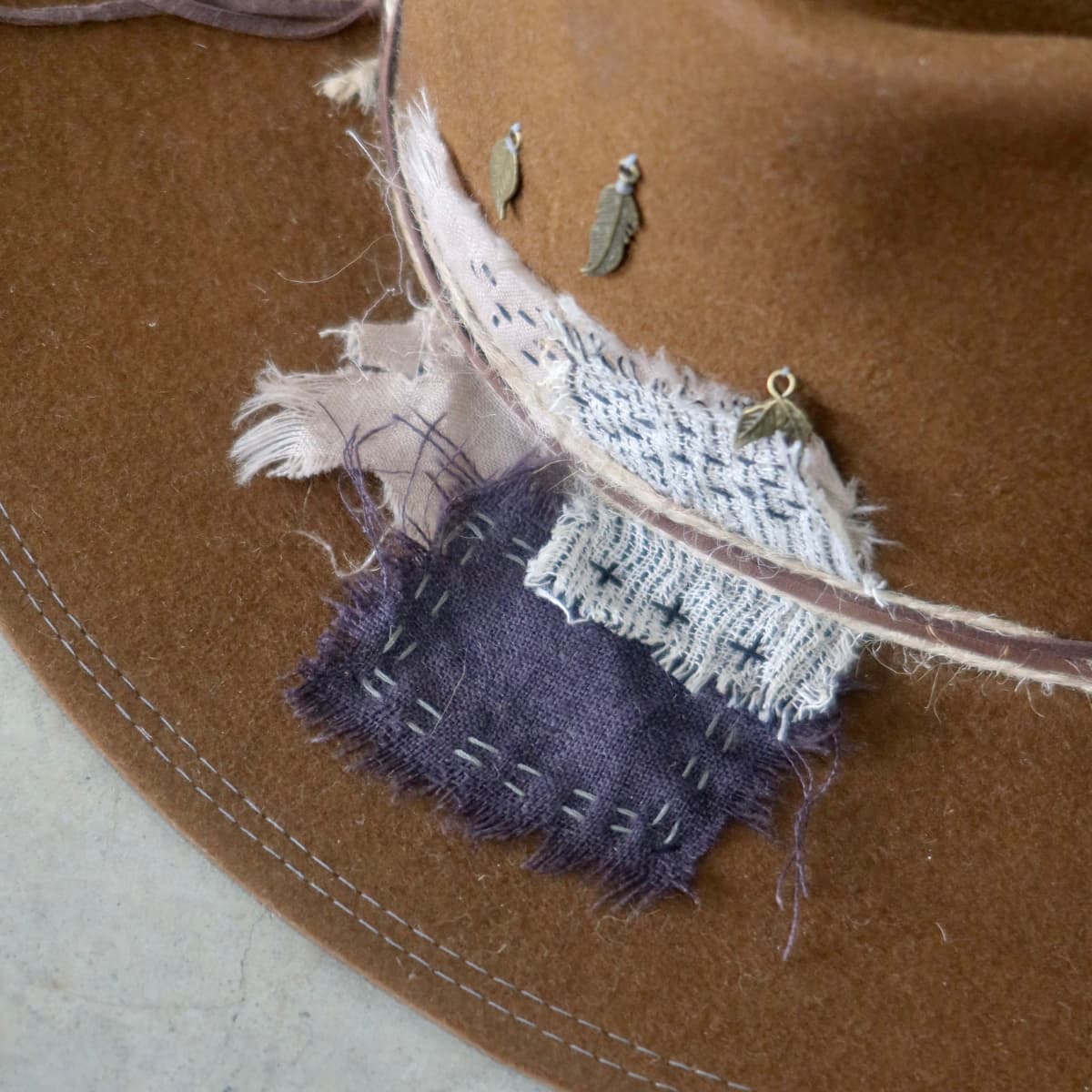 Close up detail of the George Limited Edition felt hat showing Wabi Sabi linen patchwork, hand darning stitches and antique bronze feather and leaf charms.