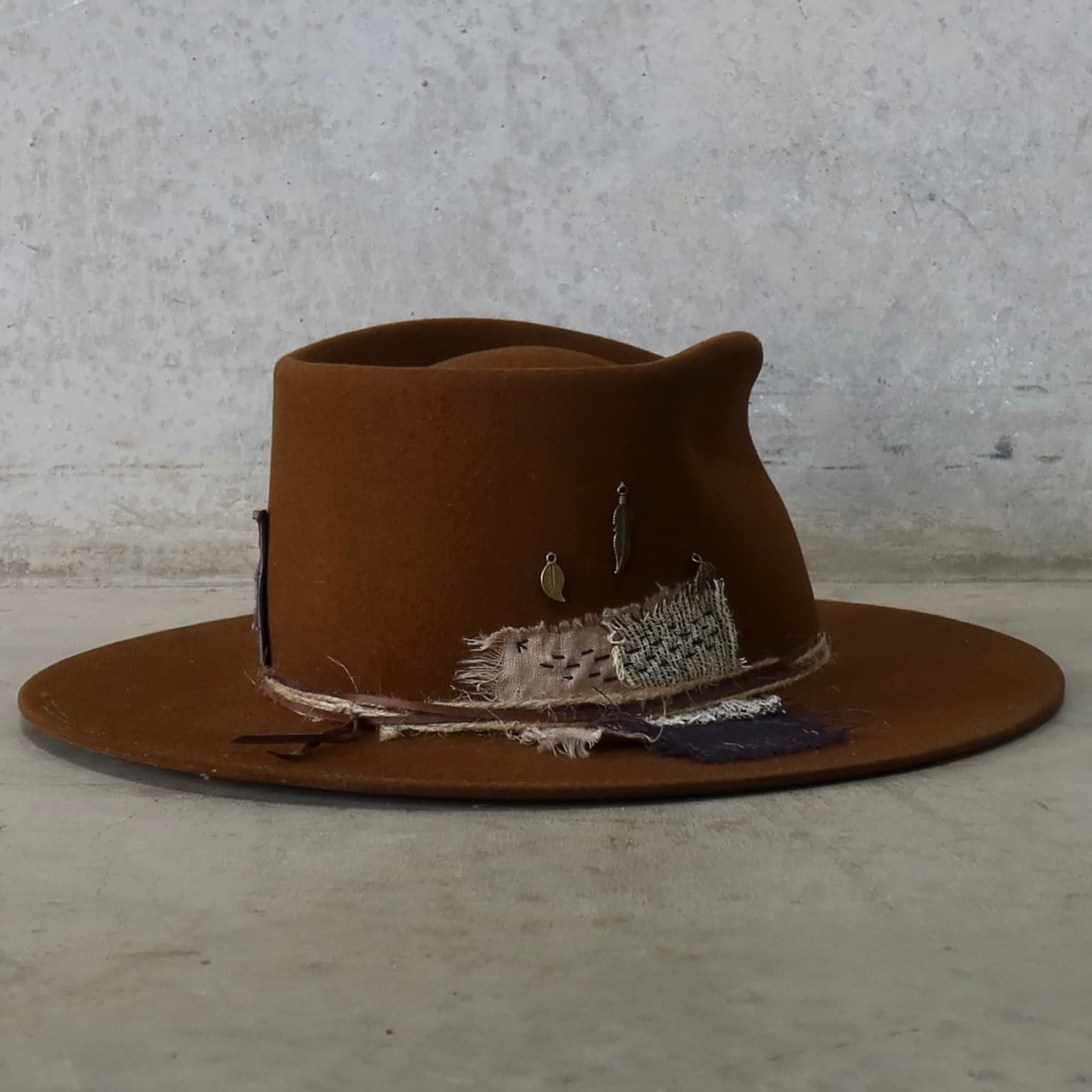 Back right angle of the caramel felt hat George Limited Edition by PHYLLi Designs featuring Wabi Sabi linen patches, antique bronze feather and leaf charms, and partial double brim stitch.