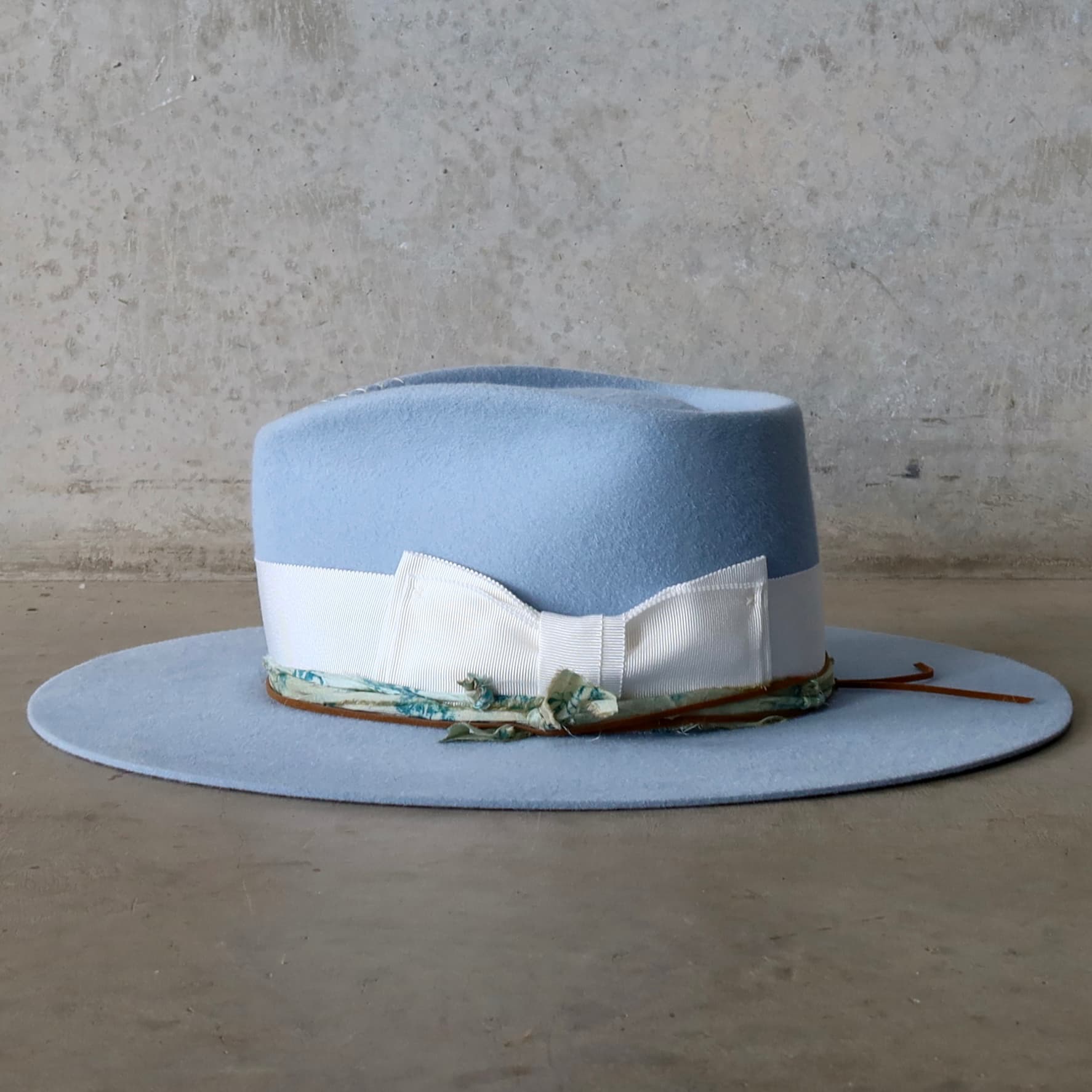 Left side view of the crystal blue felt hat Jarvis by PHYLLi Designs with ivory grosgrain bow band and blue-teal sari silk trimming.