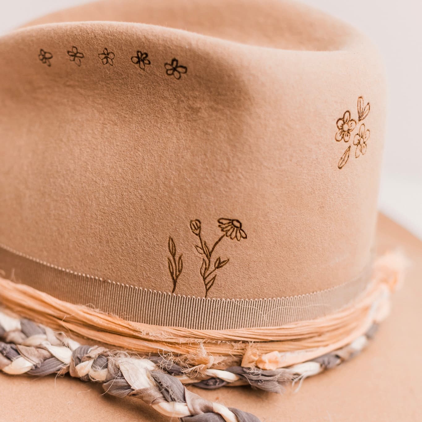 Close up detail of the La Fleurs camel felt cowboy hat by PHYLLi Designs showing burnt branded floral crown artwork above the taupe grosgrain band.