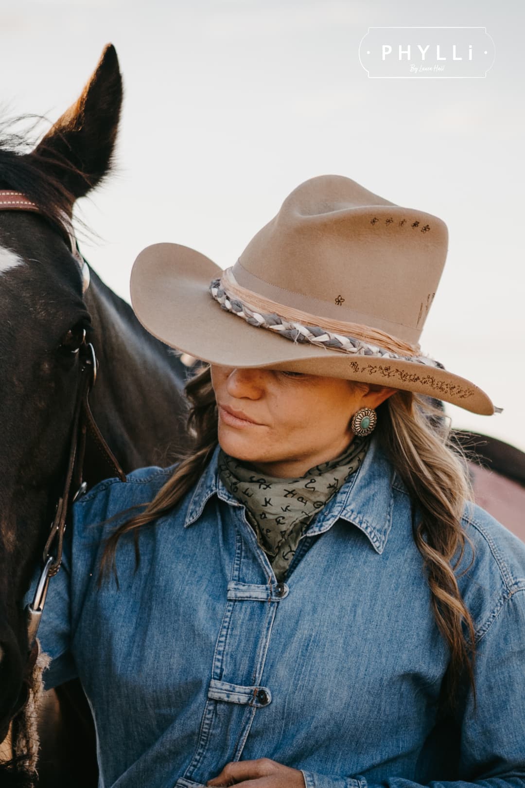 Cowgirl wearing the camel felt cowboy hat La Fleurs by PHYLLi Designs with braided sari silk trim and burnt floral branding while standing beside a saddled horse.