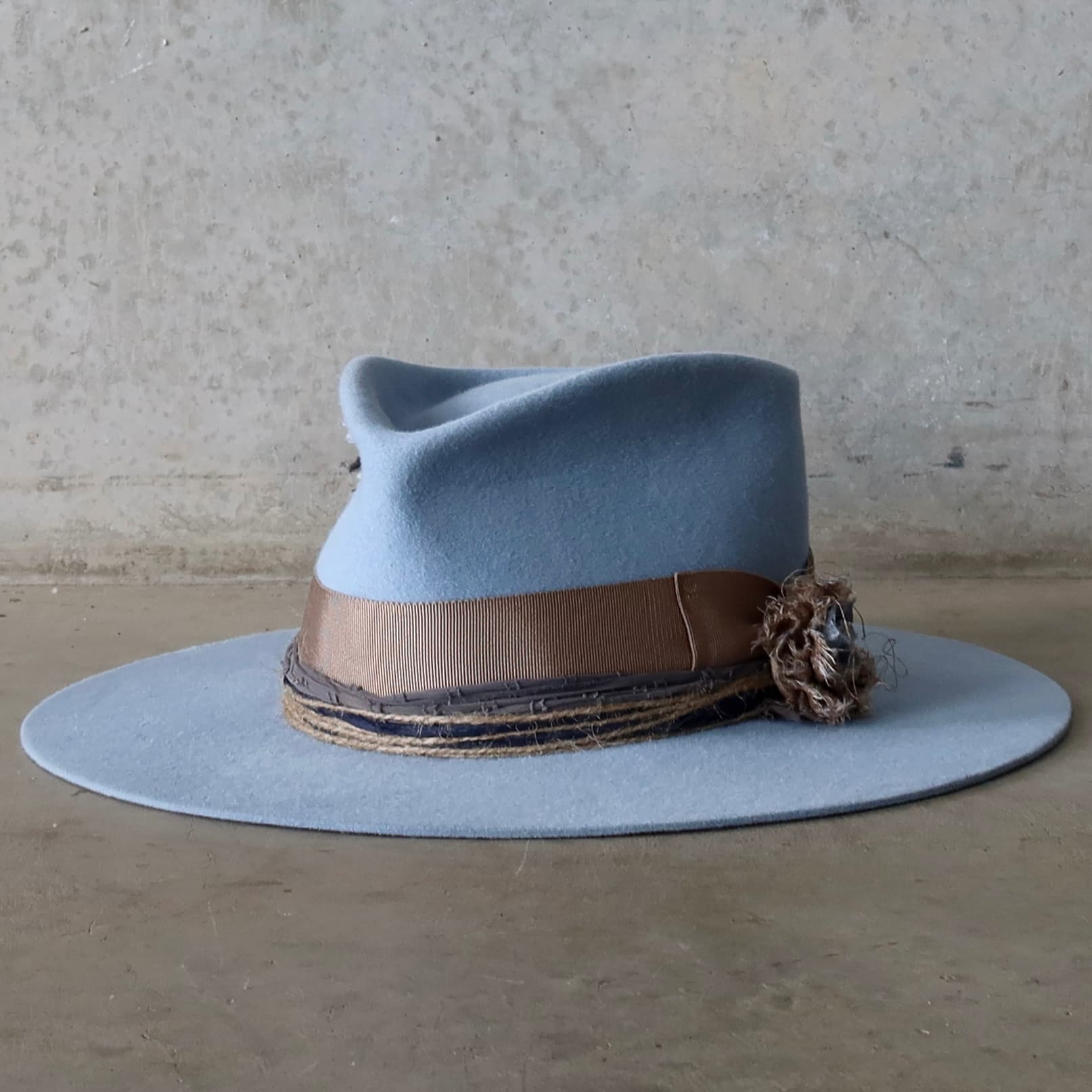 Front left angle of the crystal blue felt hat Louise by PHYLLi Designs with taupe grosgrain bow band and sari silk Lapel Bloom.