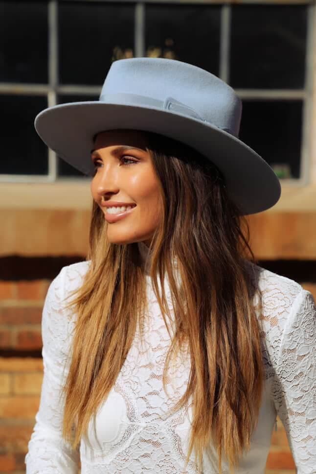 Model wearing the Crystal Blue felt hat Lucinda by PHYLLi Designs with cornflower grosgrain bow band in sunlight against a brick building.