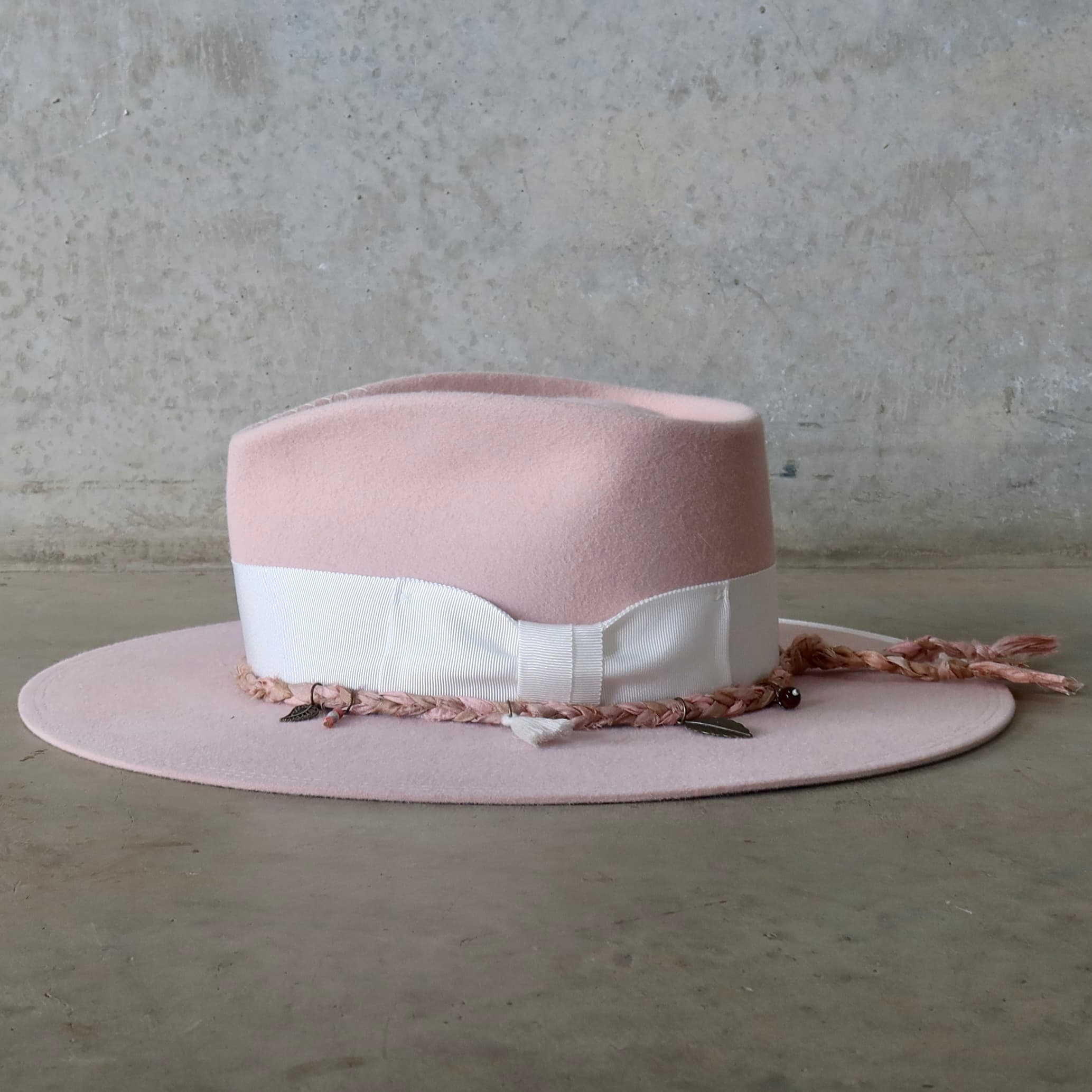 Left side view of the baby pink felt hat Lucky Charm by PHYLLi Designs with tonal pink Charm Plait featuring ivory tassel, bronze charms and bead pendants.