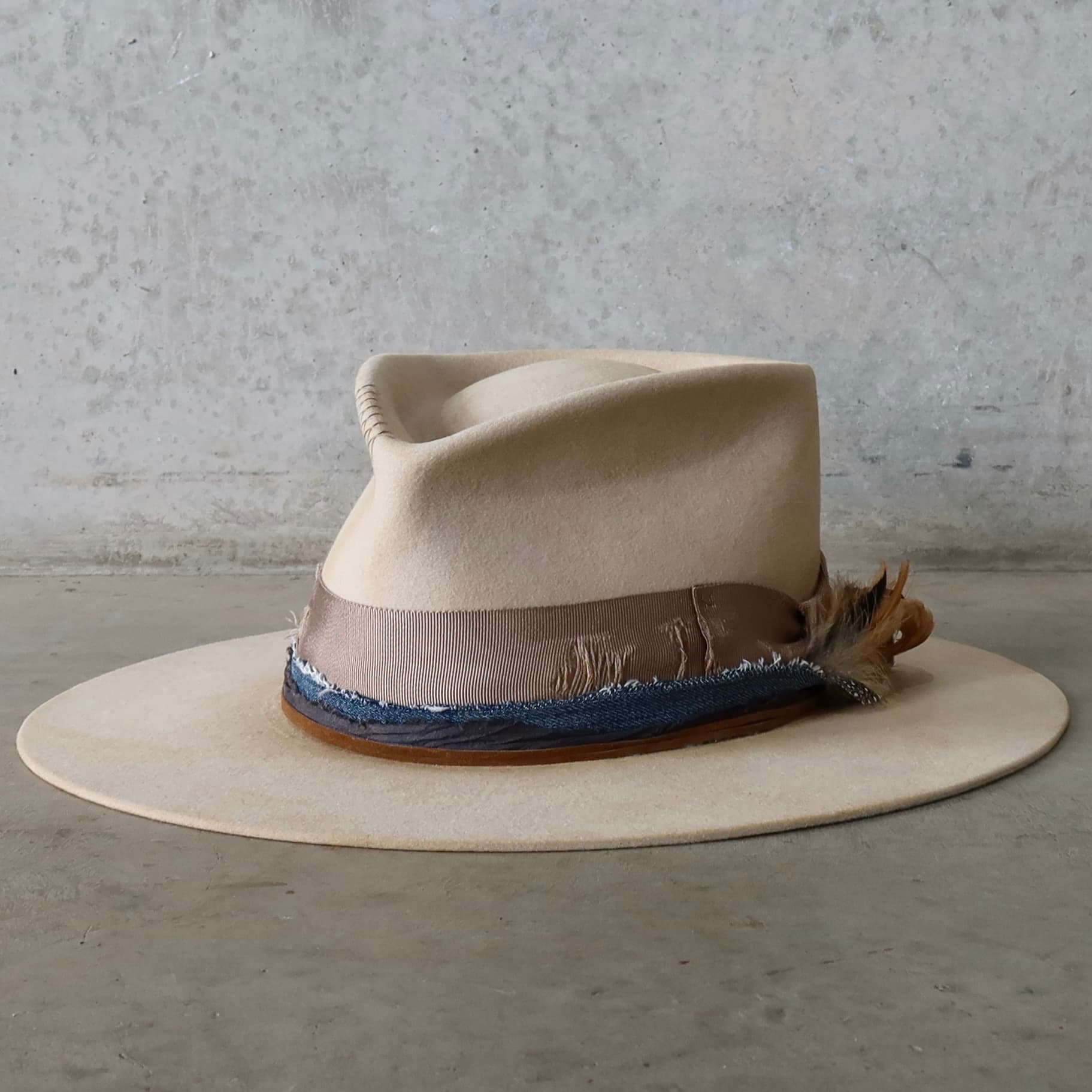 Front left angle of the beige distressed felt hat McVeigh by PHYLLi Designs with navy sari silk and denim trim, taupe grosgrain band and feather detail.