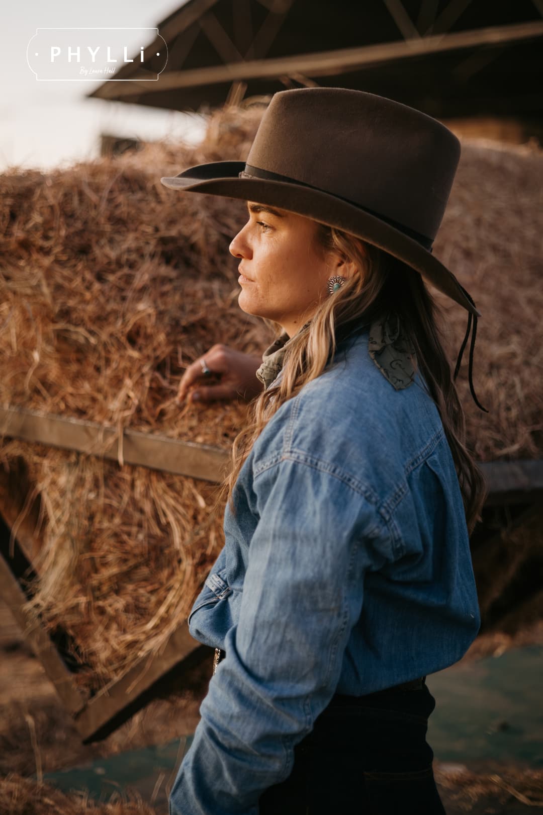 Woman wearing the Midnight Cowboy dirty olive felt cowboy hat by PHYLLi Designs with black Japanese grosgrain ribbon band in front of stacked hay bales at sunset.
