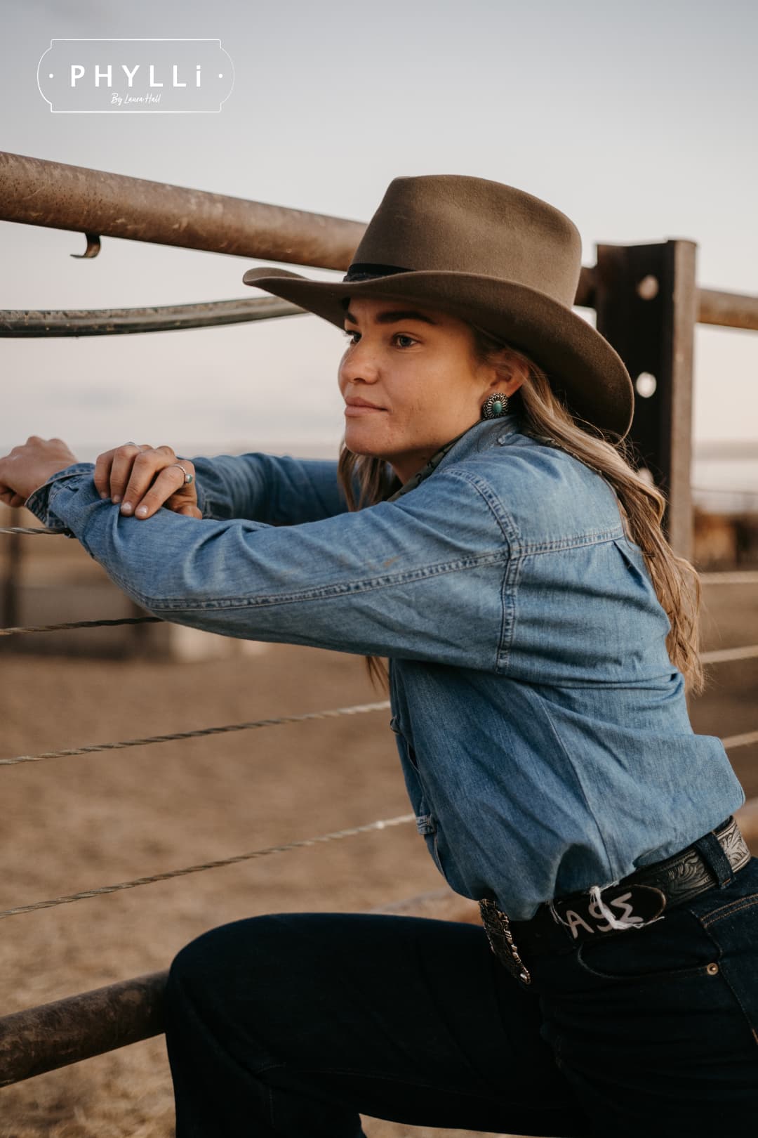 Woman wearing the Midnight Cowboy dirty olive felt cowboy hat by PHYLLi Designs with buffalo leather tieback hat band leaning on a cattle yard fence.
