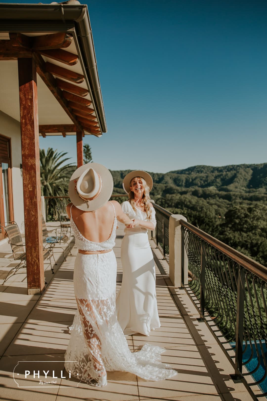 Two brides celebrating on a sunlit balcony wearing white dresses and felt bridal hats by PHYLLi Designs.