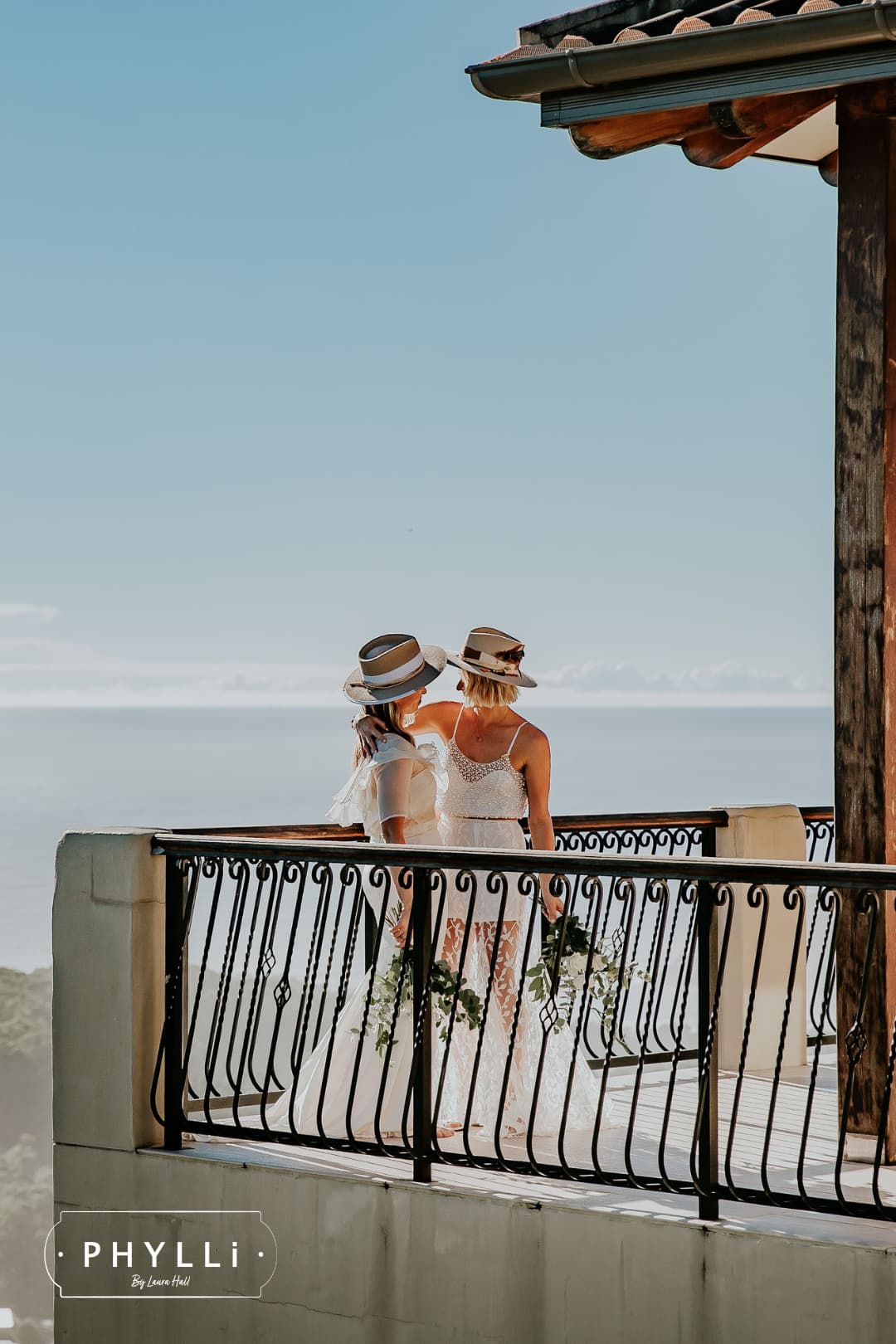 Two brides wearing statement felt wedding hats by PHYLLi Designs overlooking the ocean during a destination wedding celebration.