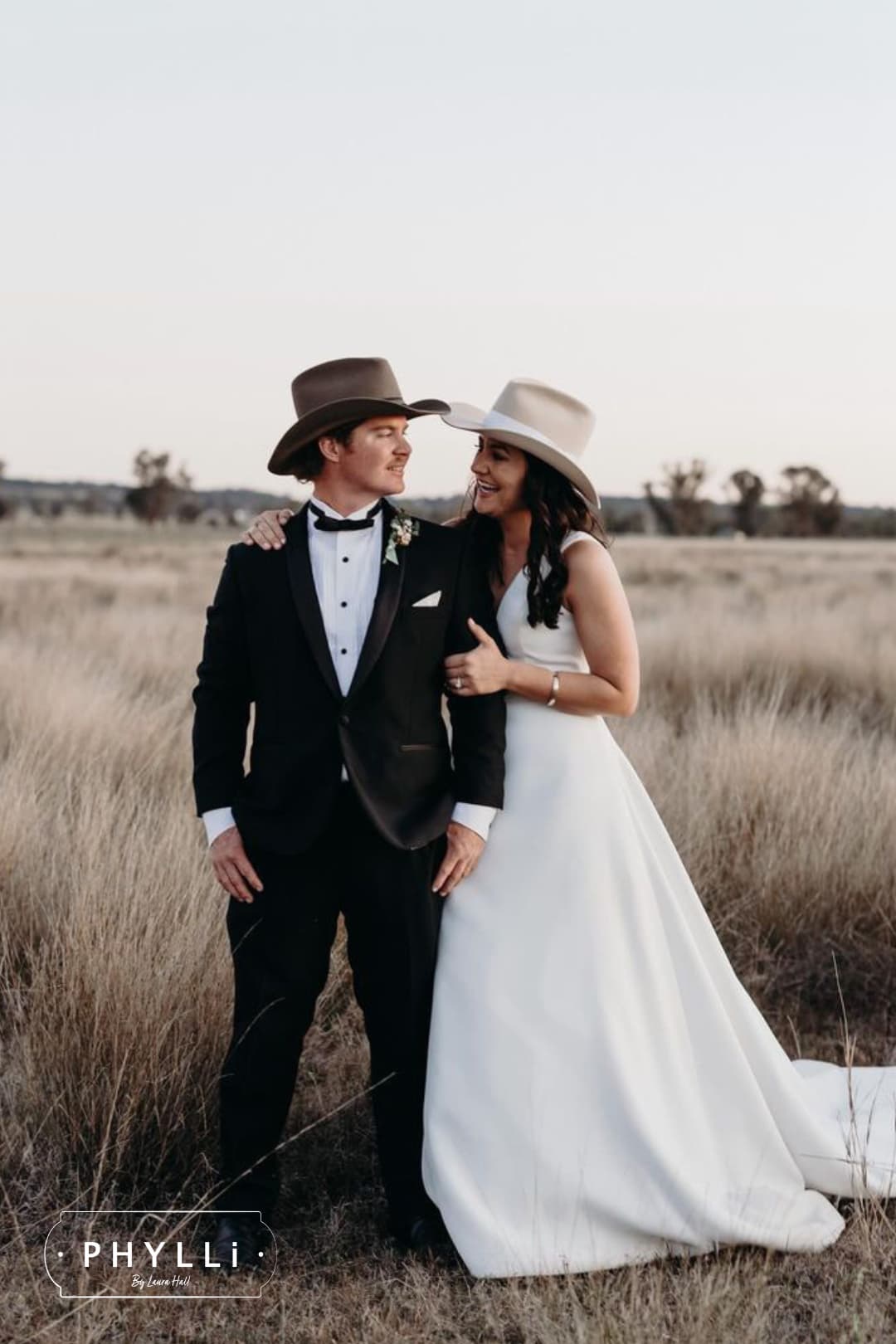 Bride and groom embracing in a rural paddock wearing felt wedding hats by PHYLLi Designs during sunset wedding portraits.