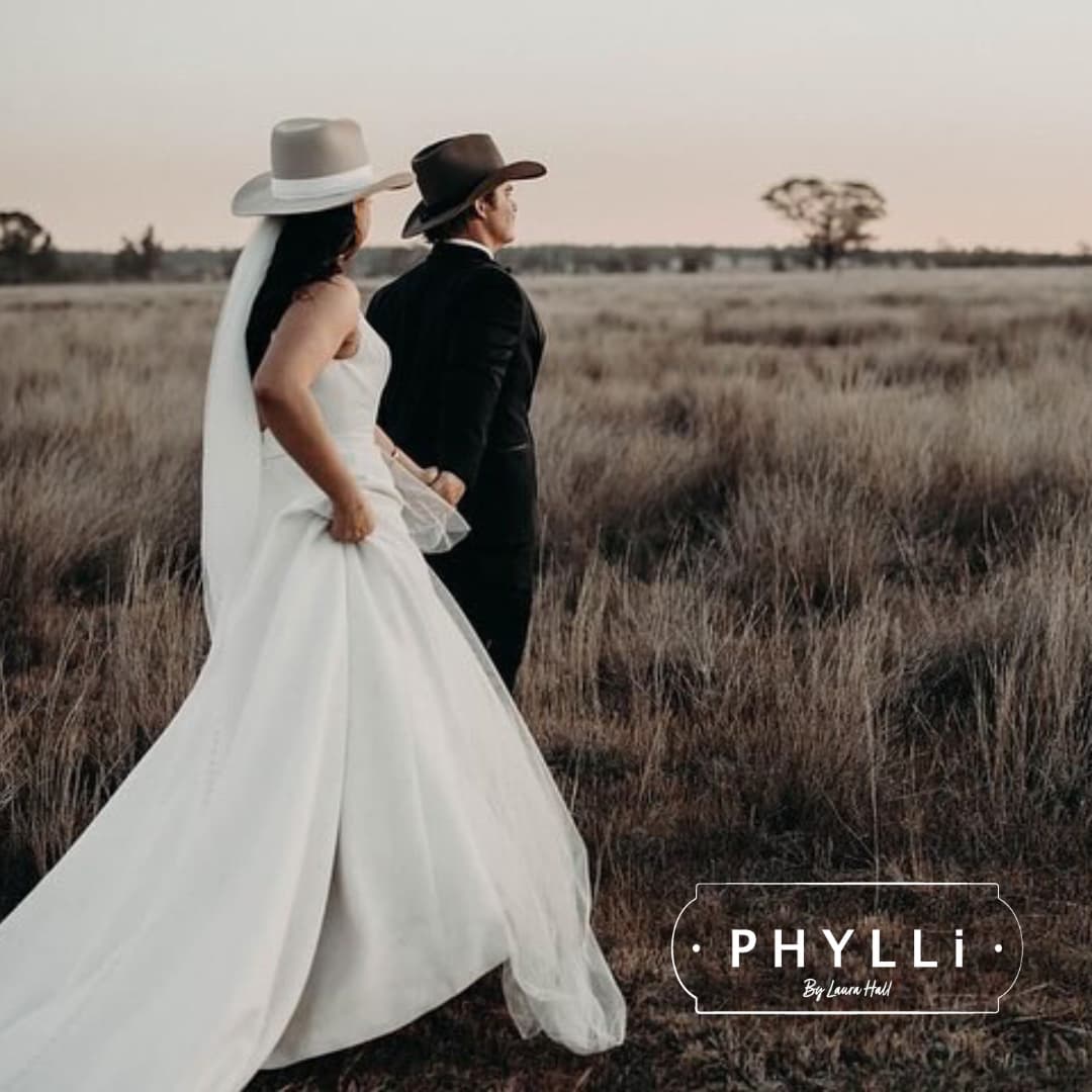 Bride and groom wearing felt wedding hats by PHYLLi Designs walking through a golden paddock during a countryside wedding photoshoot.