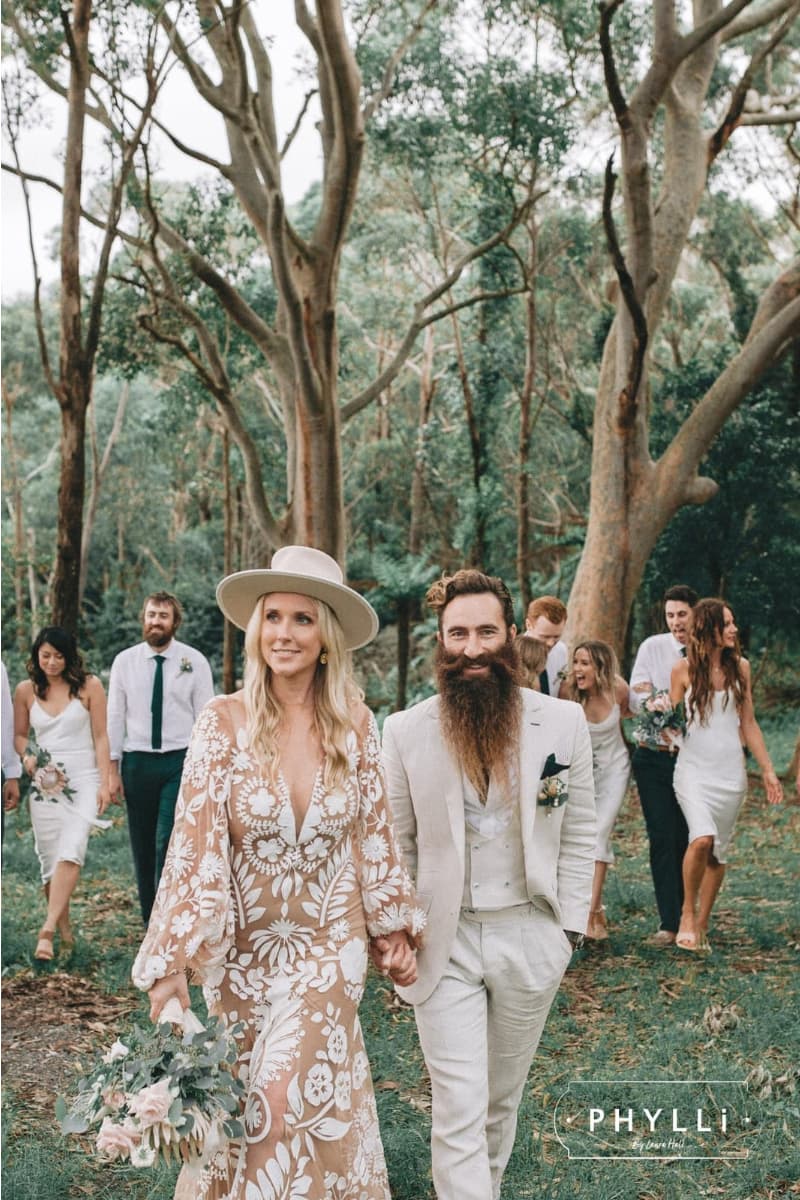 Bride and groom walking through a forest wedding setting wearing felt wedding hats by PHYLLi Designs with bridal party behind them.