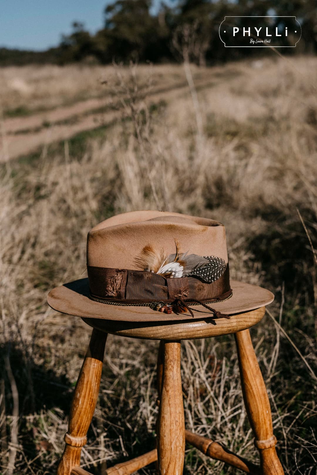 Roadie distressed camel felt hat by PHYLLi Designs displayed on a wooden stool outdoors.