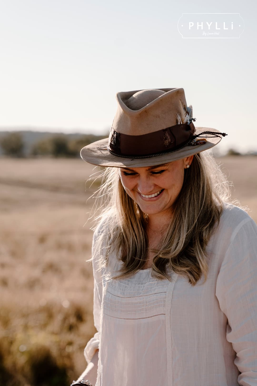 Laura Hall wearing the Roadie distressed camel felt hat by PHYLLi Designs in a rural field.