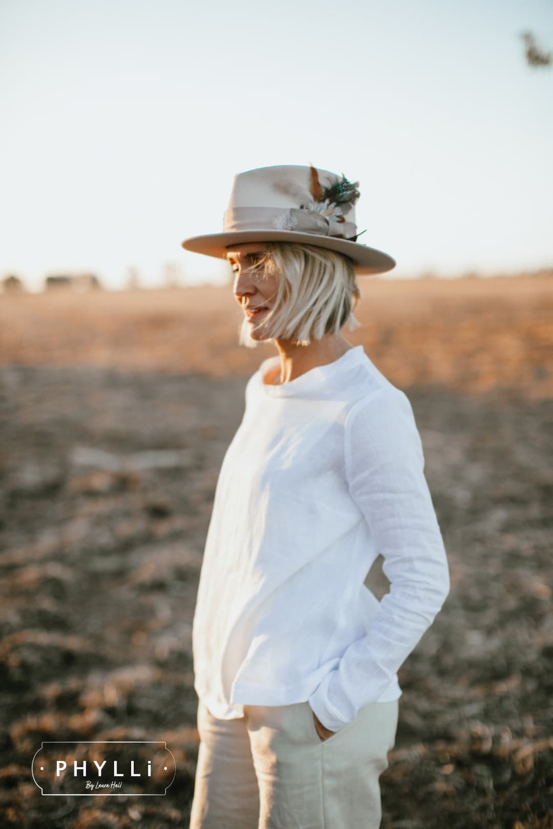 Woman wearing the Roadie beige felt hat by PHYLLi Designs with feather cluster and distressed grosgrain band in a dry rural field at sunset.