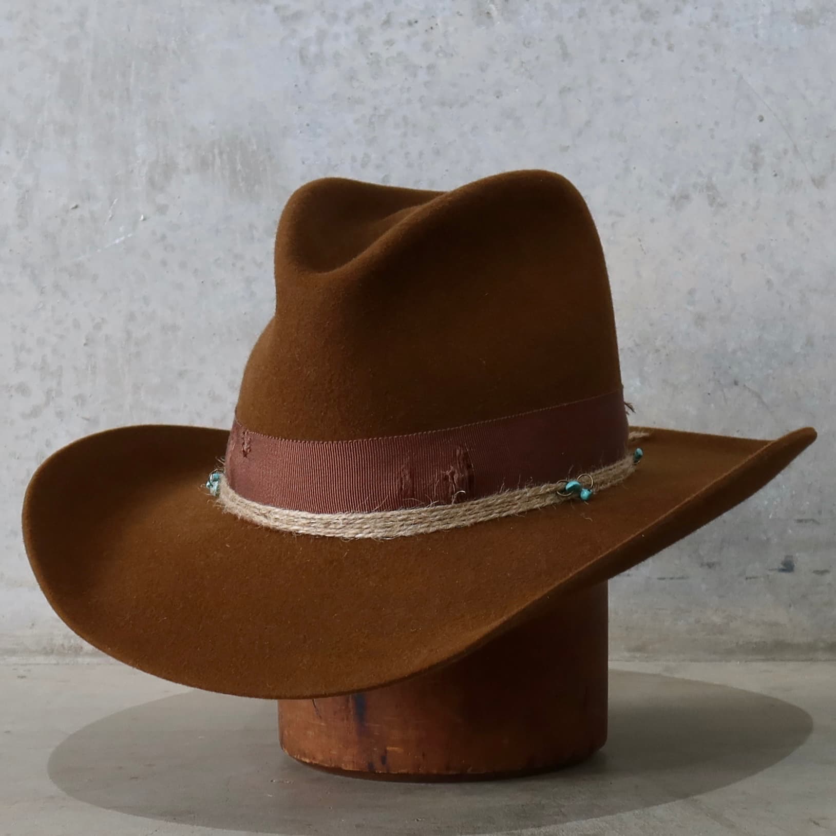 Front left angle of the caramel cowboy hat Rue by PHYLLi Designs with distressed chocolate grosgrain band and turquoise beaded twine wraps.