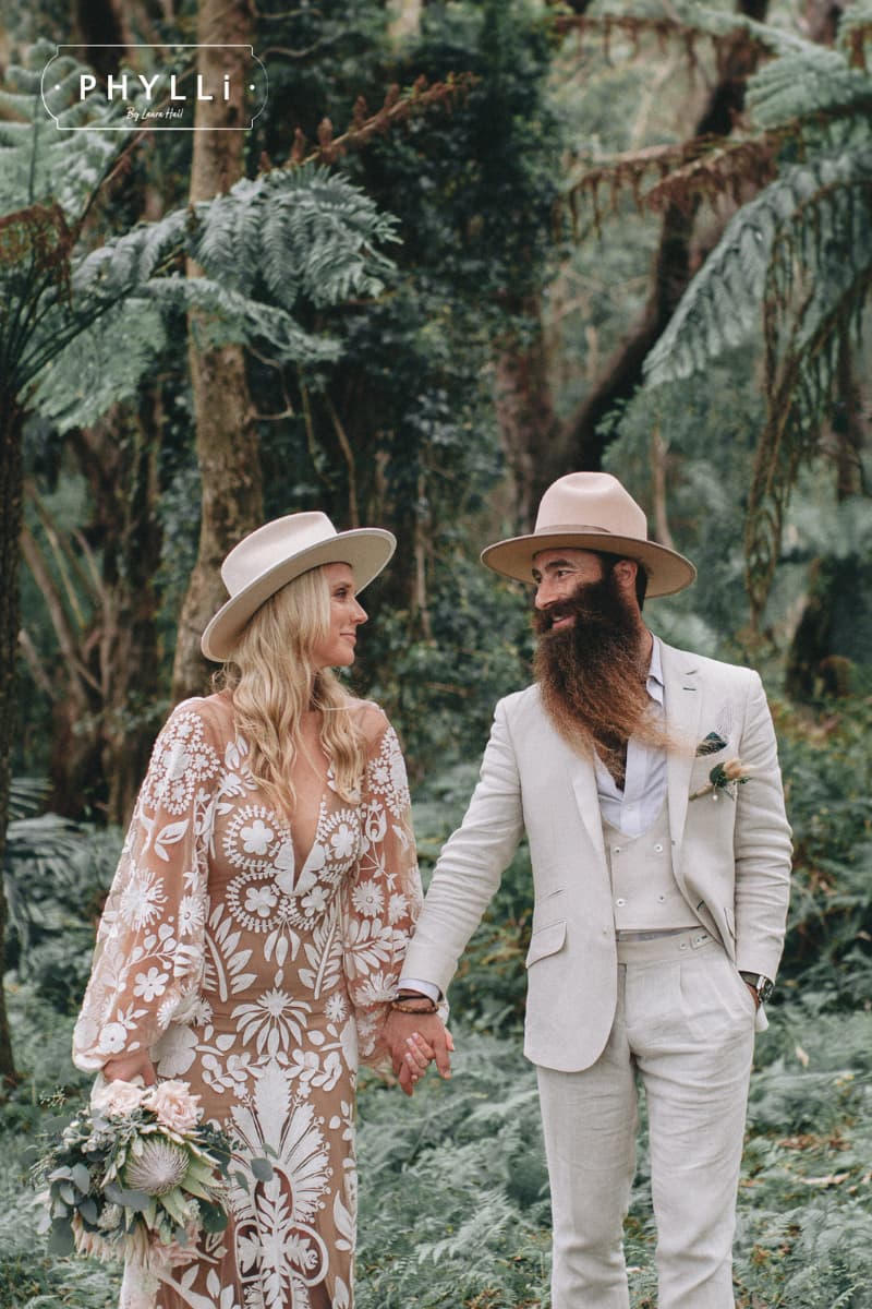 Bride wearing the Rue De Seine beige felt hat by PHYLLi Designs with ivory grosgrain bow band and silver hat pin with groom in a forest wedding setting.