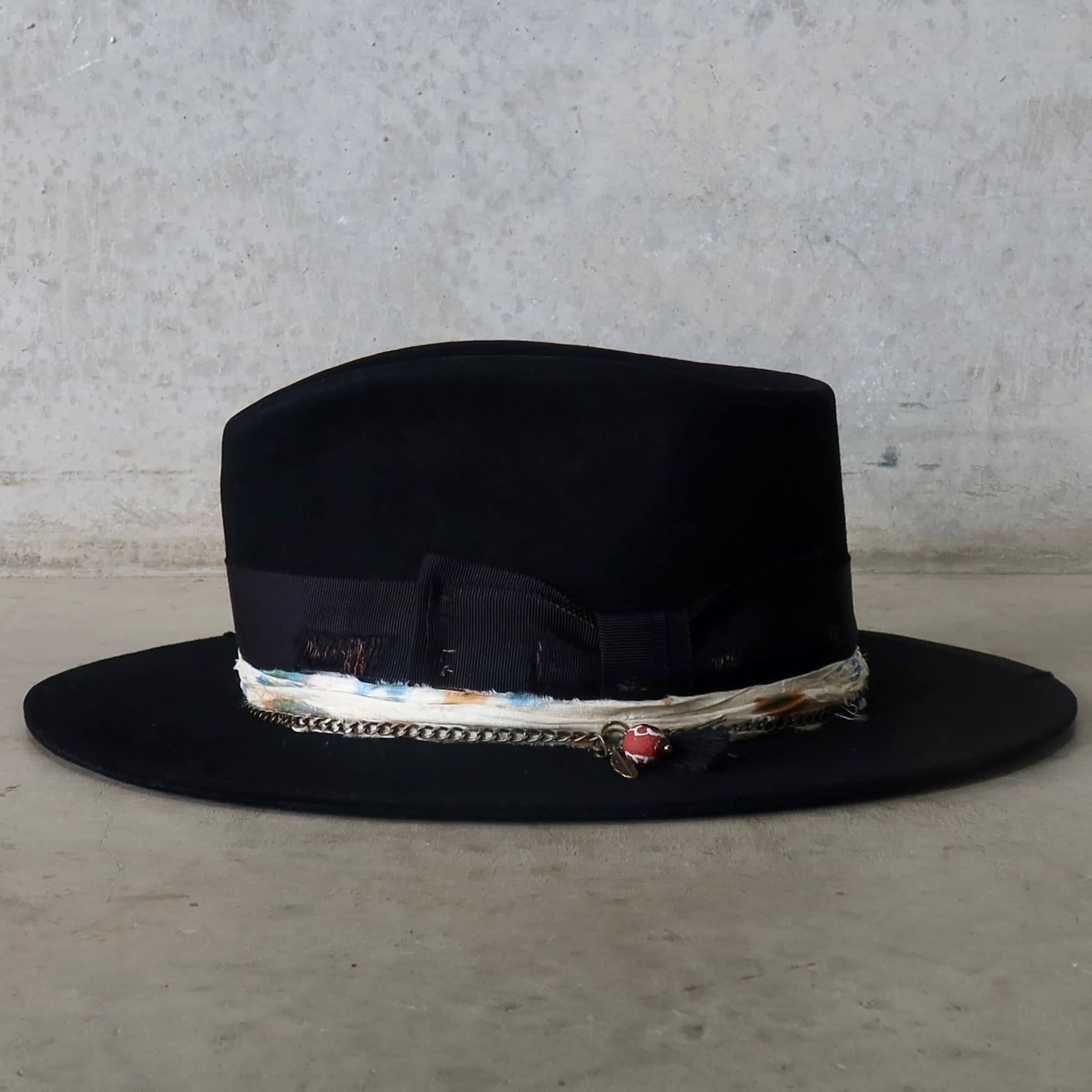 Left side view of the black felt hat Saint by PHYLLi Designs featuring distressed Japanese grosgrain double bow band and charm cluster with black tassel, bronze charms and recycled glass Ghana bead pendant.