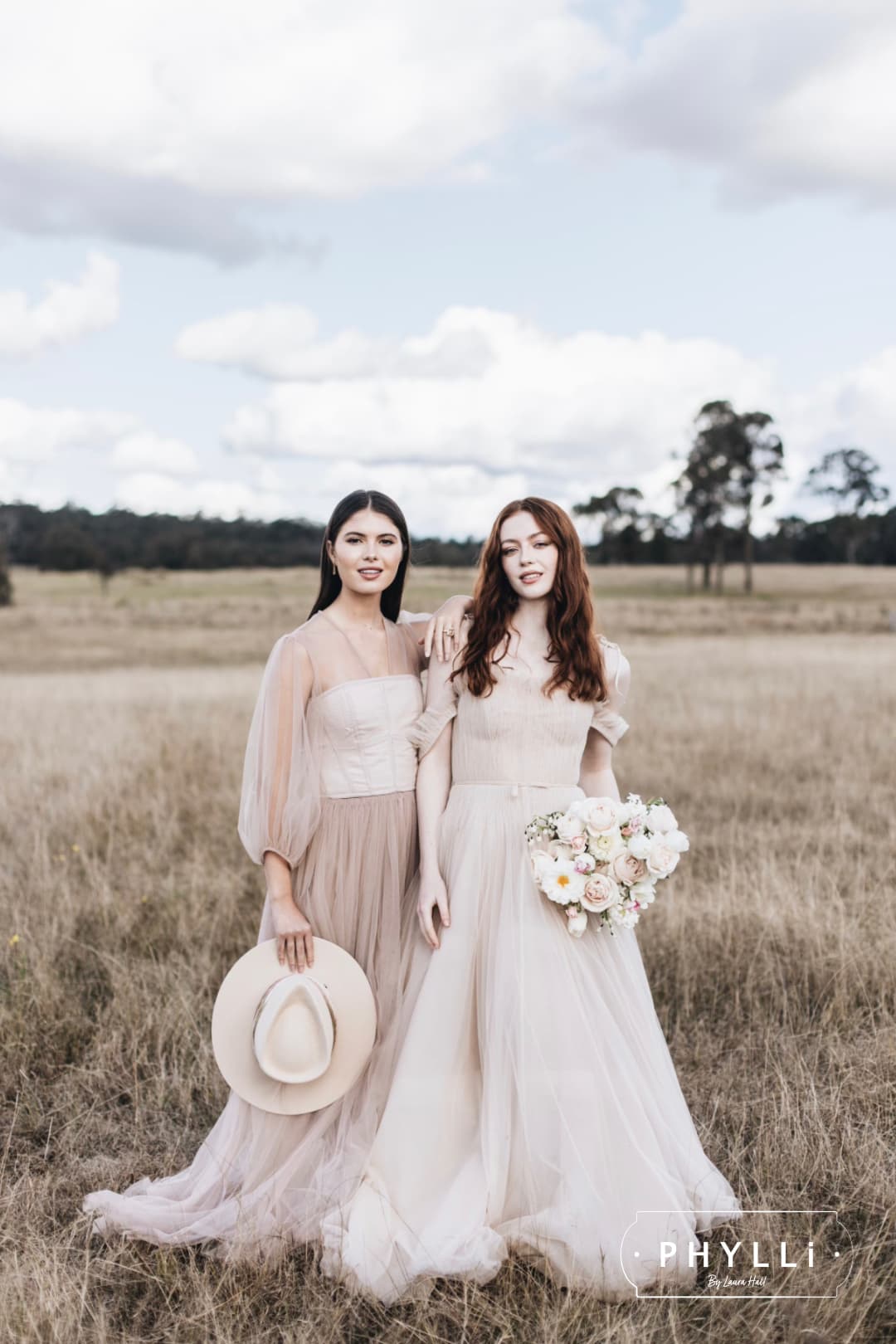 Two brides in wedding dresses standing in a field, one holding a beige felt hat by PHYLLi Designs.