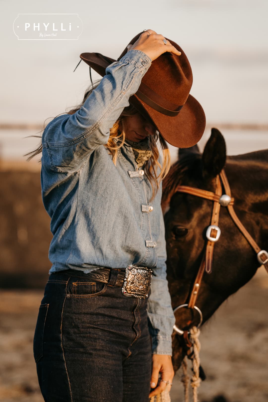 Woman wearing the chocolate cowboy hat Spicer by PHYLLi Designs with chocolate grosgrain band adjusting her hat beside a bridled horse in an outback paddock.