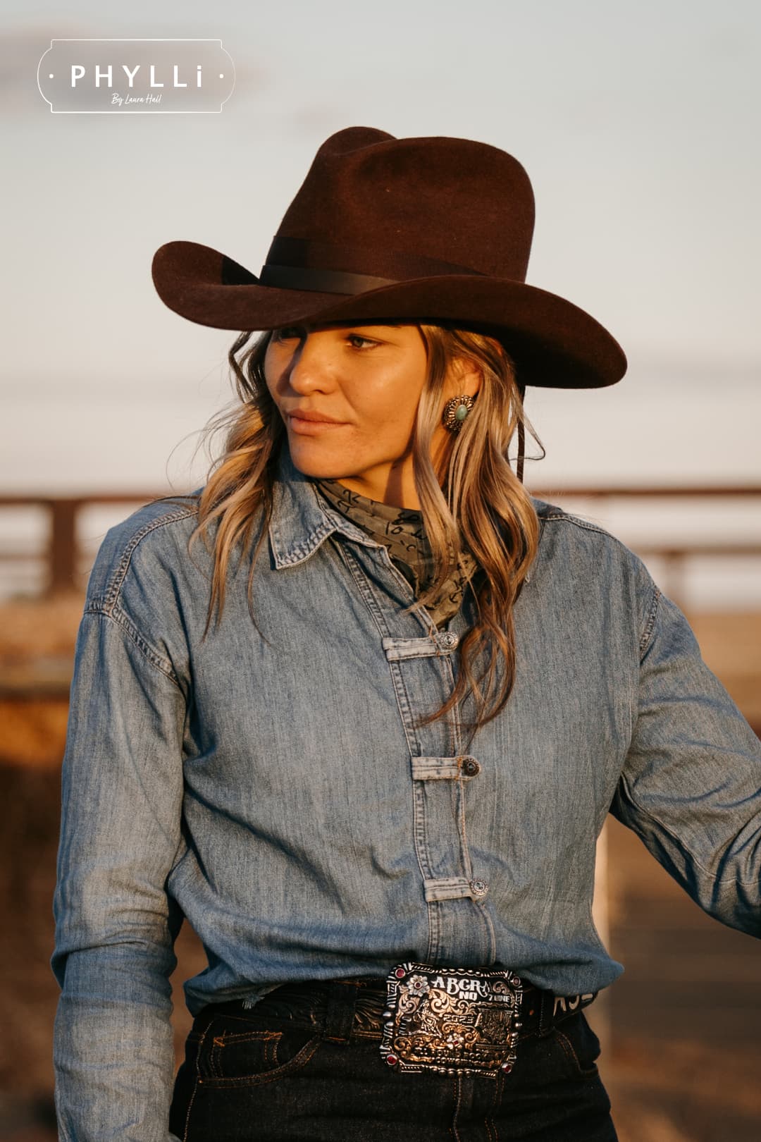 Model wearing the chocolate cowboy hat Spicer by PHYLLi Designs with chocolate grosgrain band and leather tie back in a rural paddock at sunset.