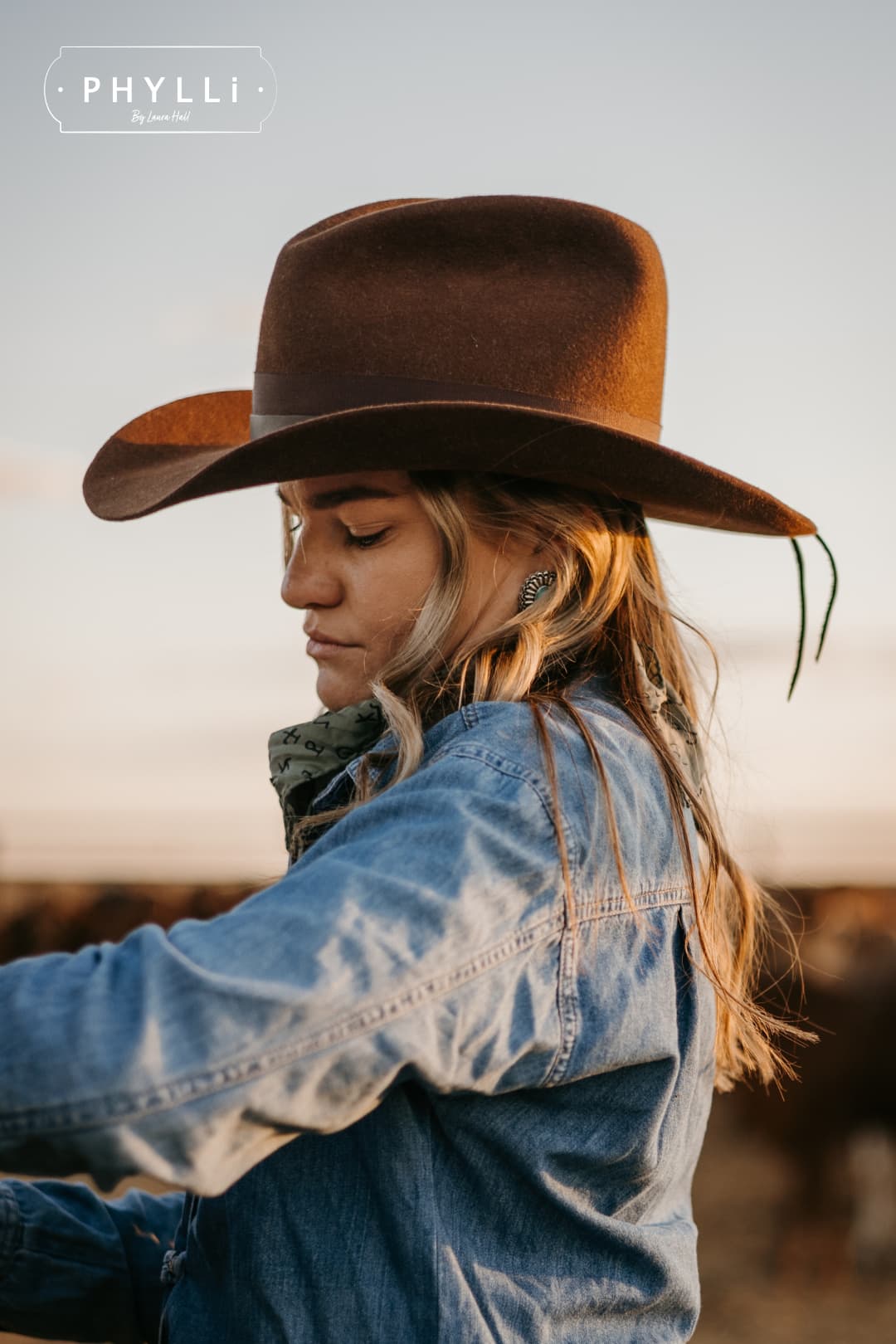 Woman wearing the chocolate cowboy hat Spicer by PHYLLi Designs with leather tie back band in an outback paddock beside a cattle.