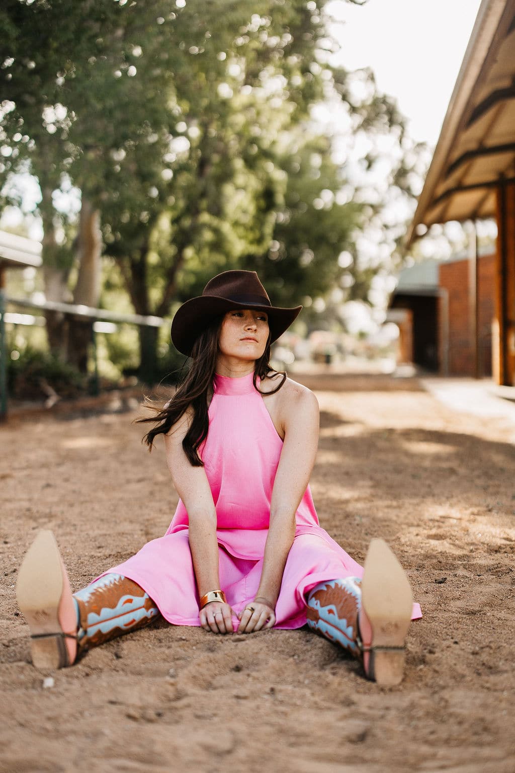 Woman wearing the PHYLLi Spicer chocolate cowboy hat with a pink dress and cowboy boots, sitting on the ground by a stable.