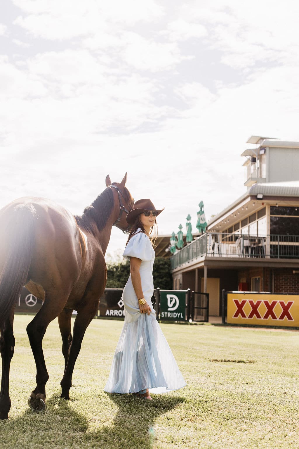 Woman wearing the PHYLLi Spicer cowboy hat with a pale blue dress, standing beside a horse trackside at a racecourse.