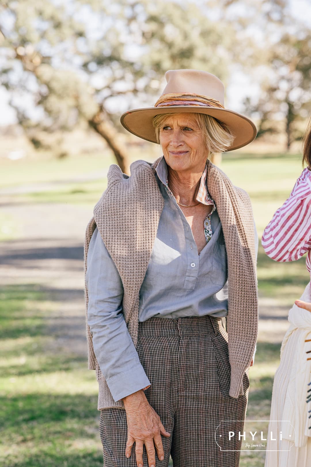 Older woman wearing the Sunset sand felt hat by PHYLLi Designs with a knit draped over her shoulders in a sunlit rural setting.