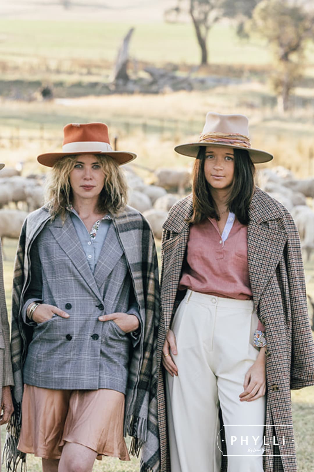 Two women standing outdoors in tailored layers, with the woman on the right wearing the Sunset sand felt hat by PHYLLi Designs.