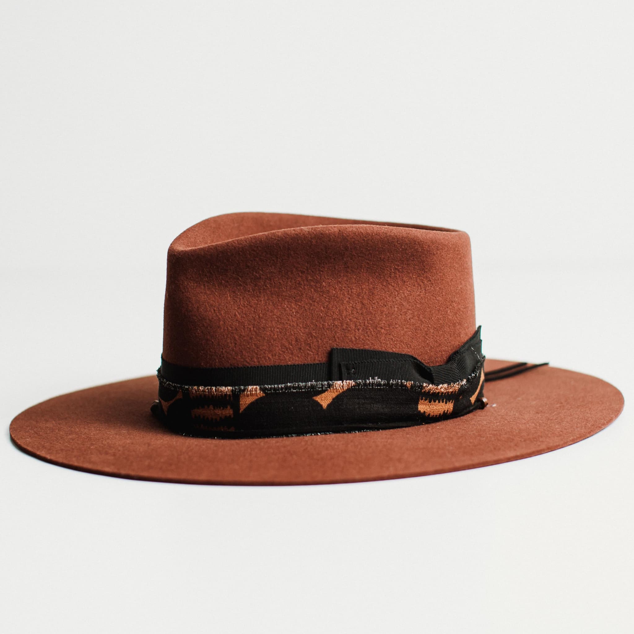 Angled view of russet felt hat with decorative band by PHYLLi Designs on a light grey background.