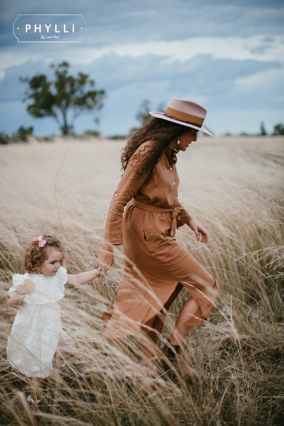 Woman wearing The Blush felt hat by PHYLLi Designs walking through tall grass holding child’s hand.
