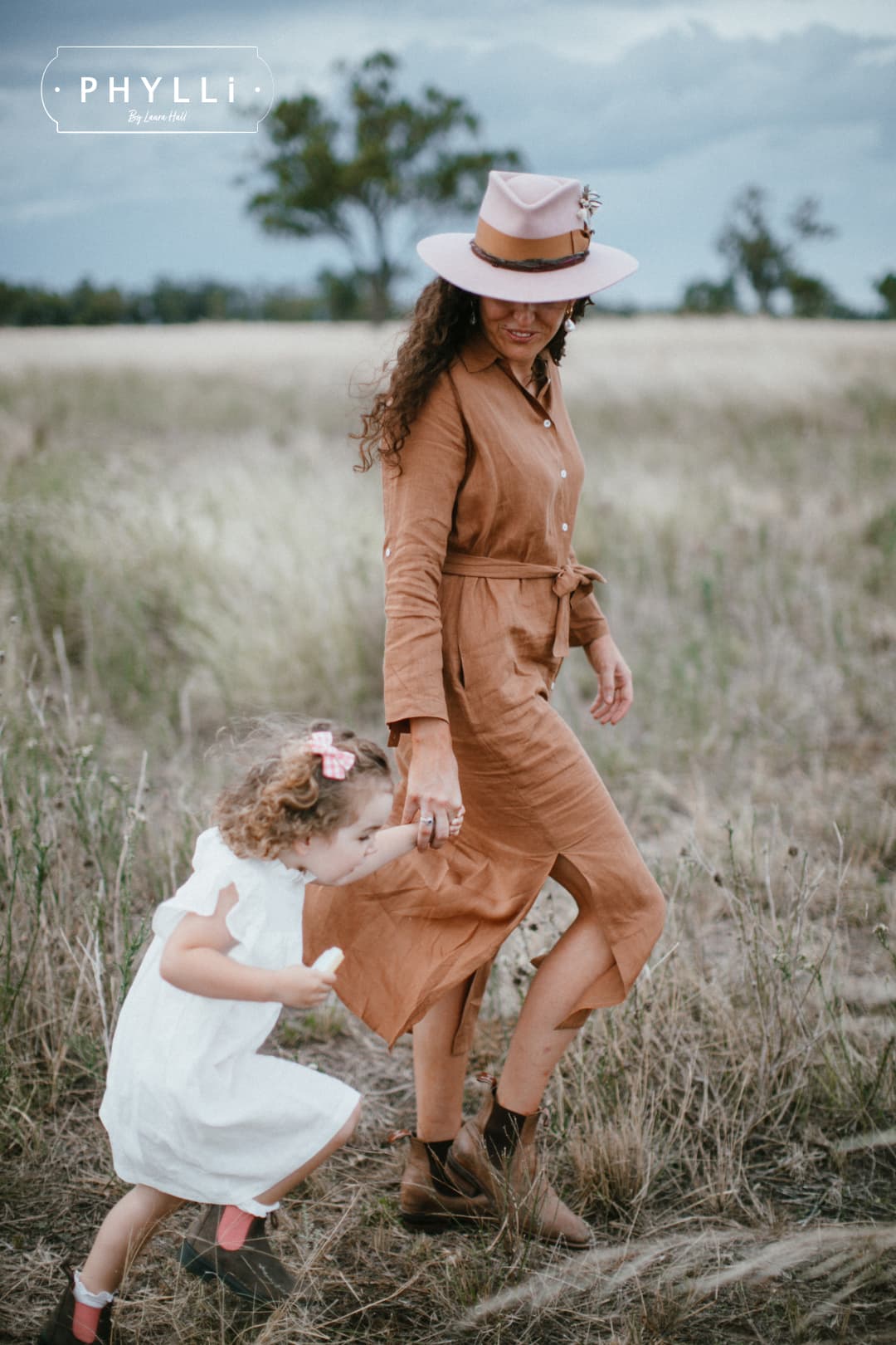 Mother wearing The Blush felt hat walking through field holding child’s hand.